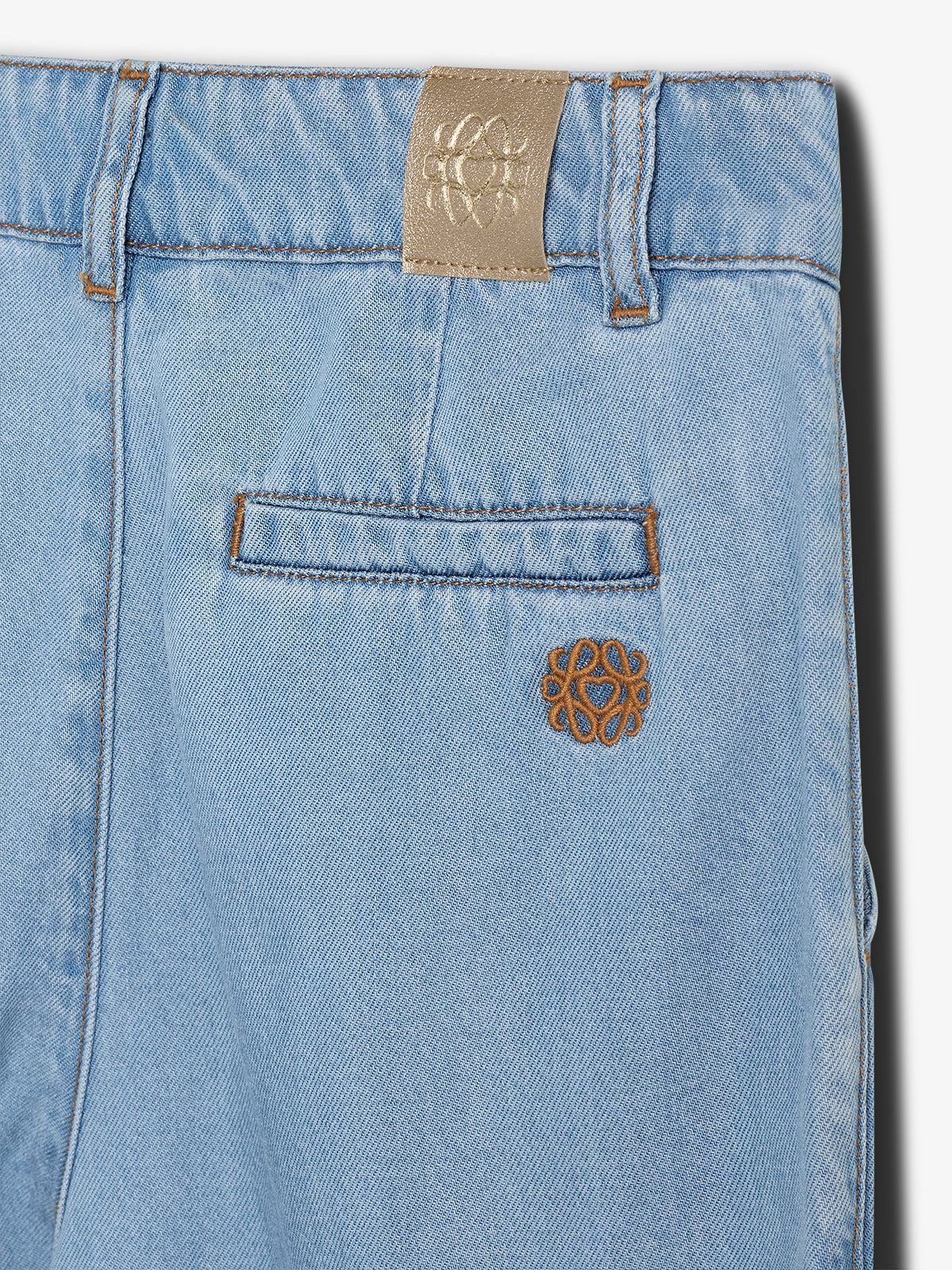 Lyseblå denimshorts med en lille baglomme, markeret af brun syning. Der er et beige mærke med et symmetrisk logo ved taljen.