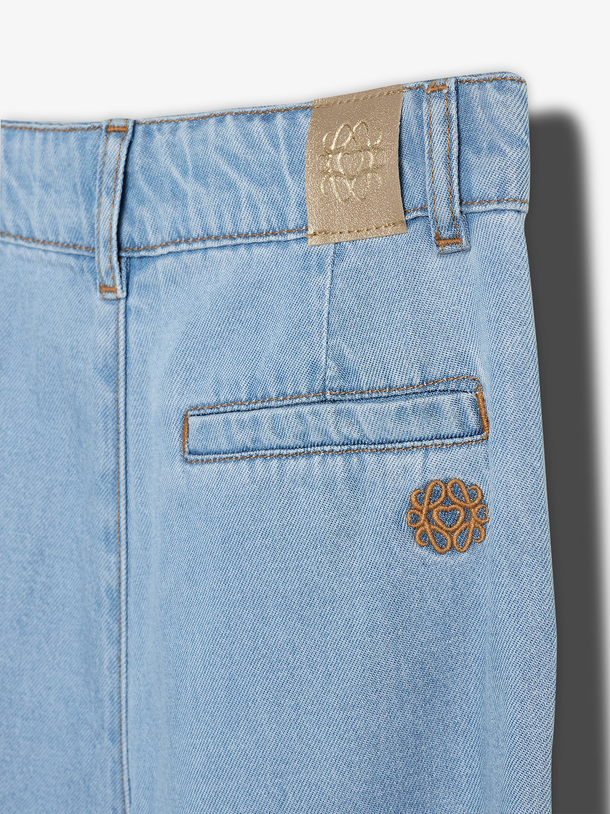 Light blue denim pants with brown stitching and embroidered design, featuring a stitched logo patch at the waistband and small decorative emblem near the pocket, against a plain background.