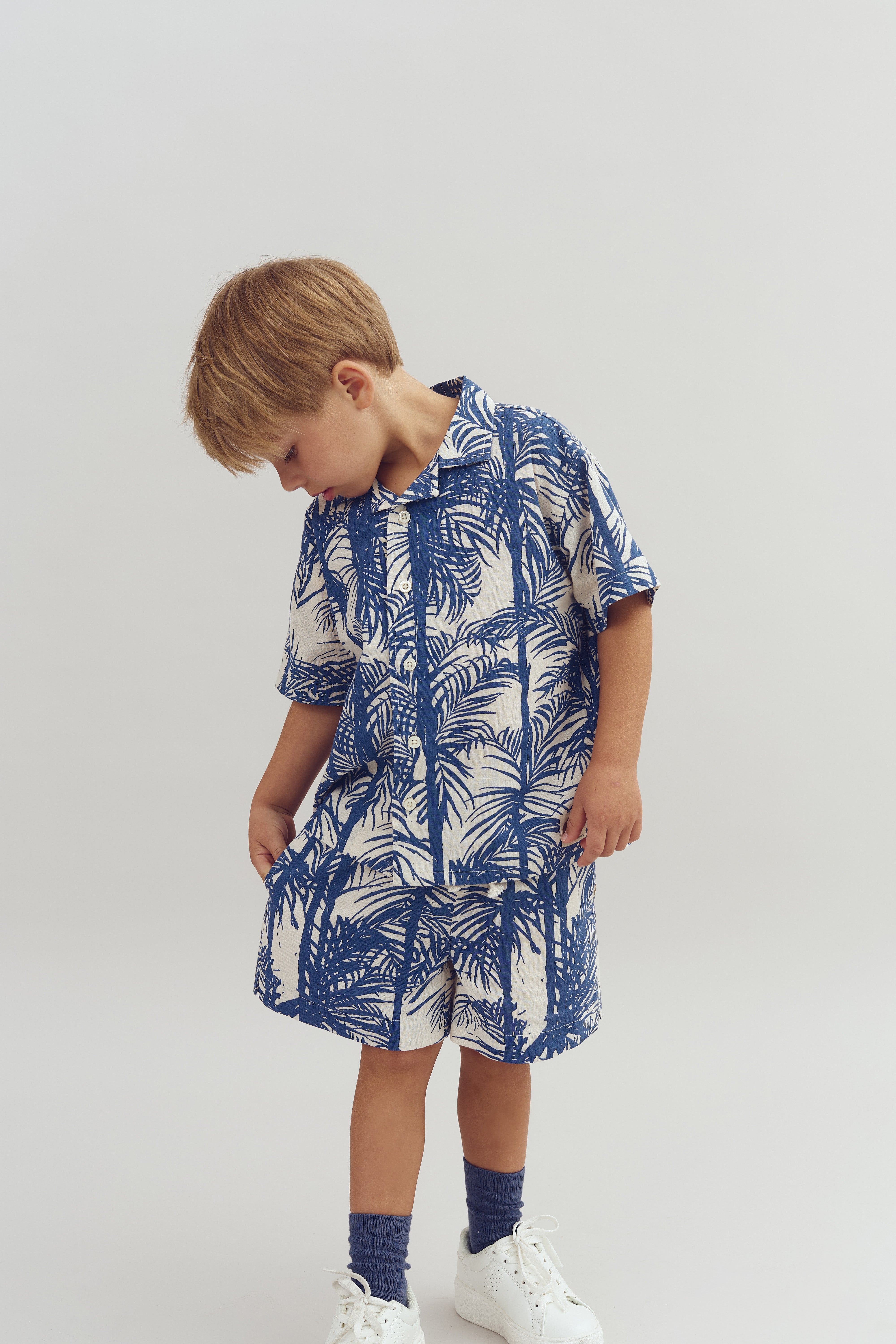 A young child stands, looking down, wearing a blue and white tropical-print outfit. The child has light brown hair and white sneakers, set against a plain, light background.