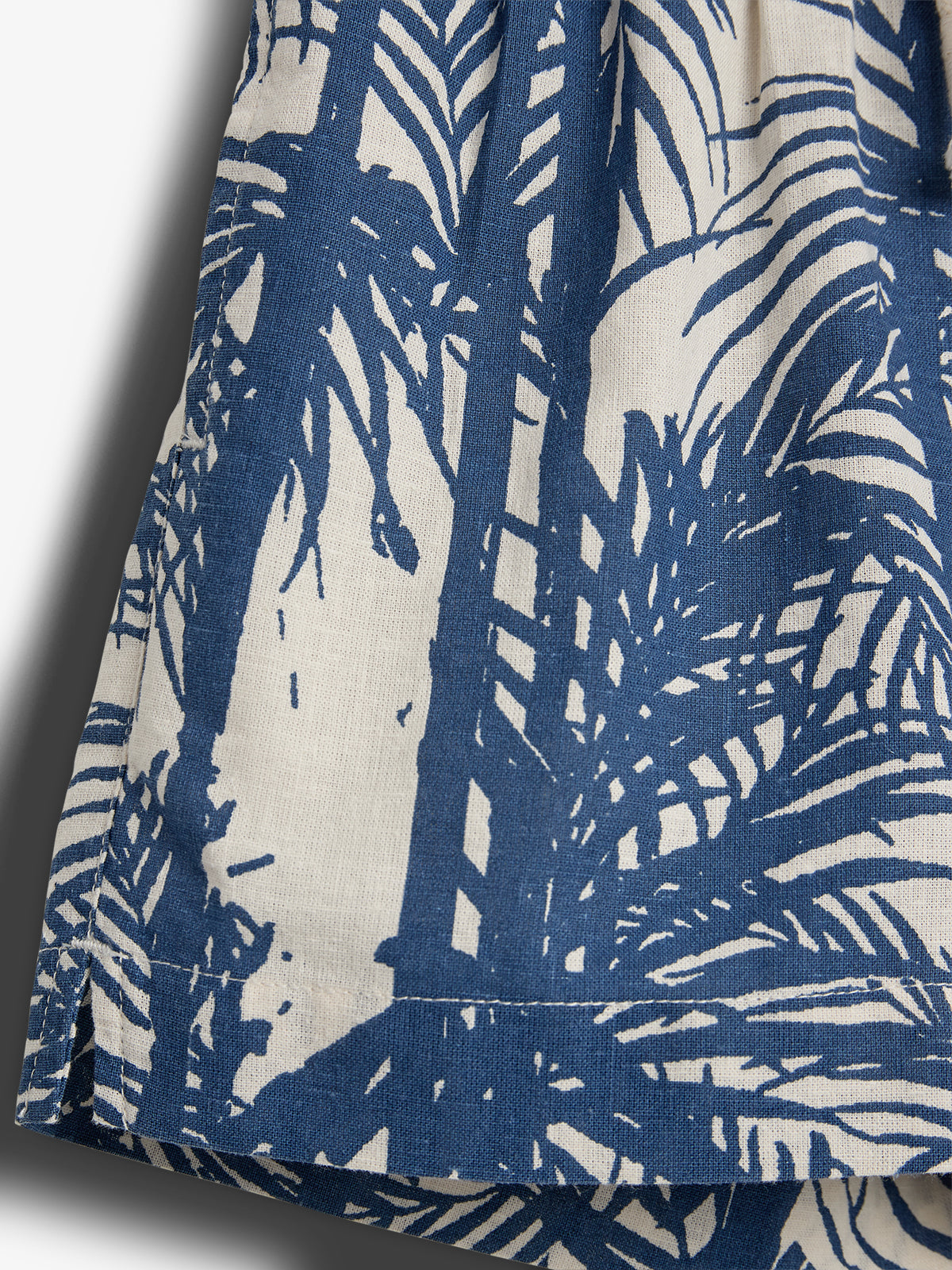 Fabric with a blue palm leaf pattern on a cream background, folded and slightly gathered, casting a shadow on a plain surface.