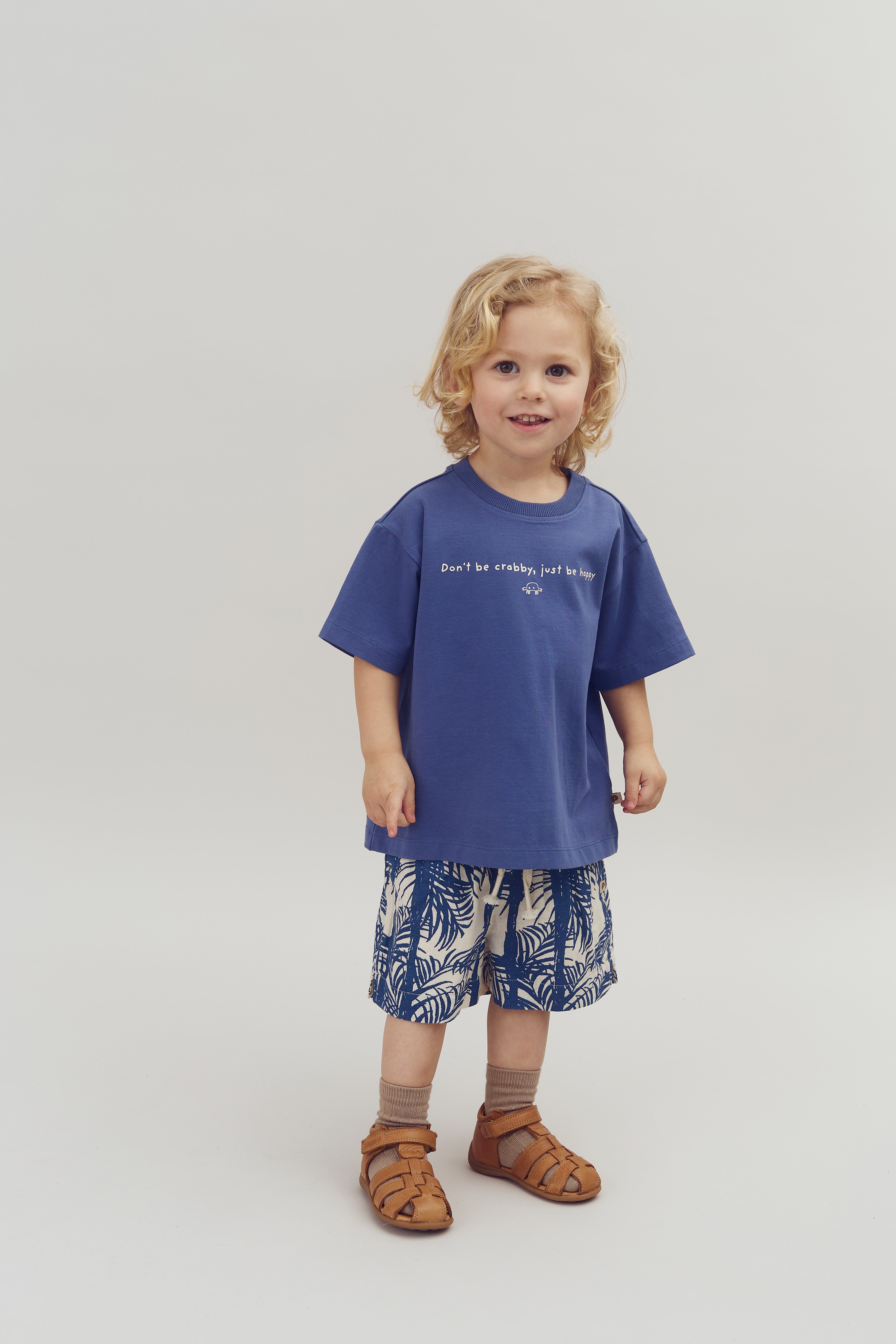 Child stands smiling in a studio, wearing a blue shirt with text: "Don't be crabby, just be happy," and beige shorts with blue leaf patterns, paired with brown sandals.