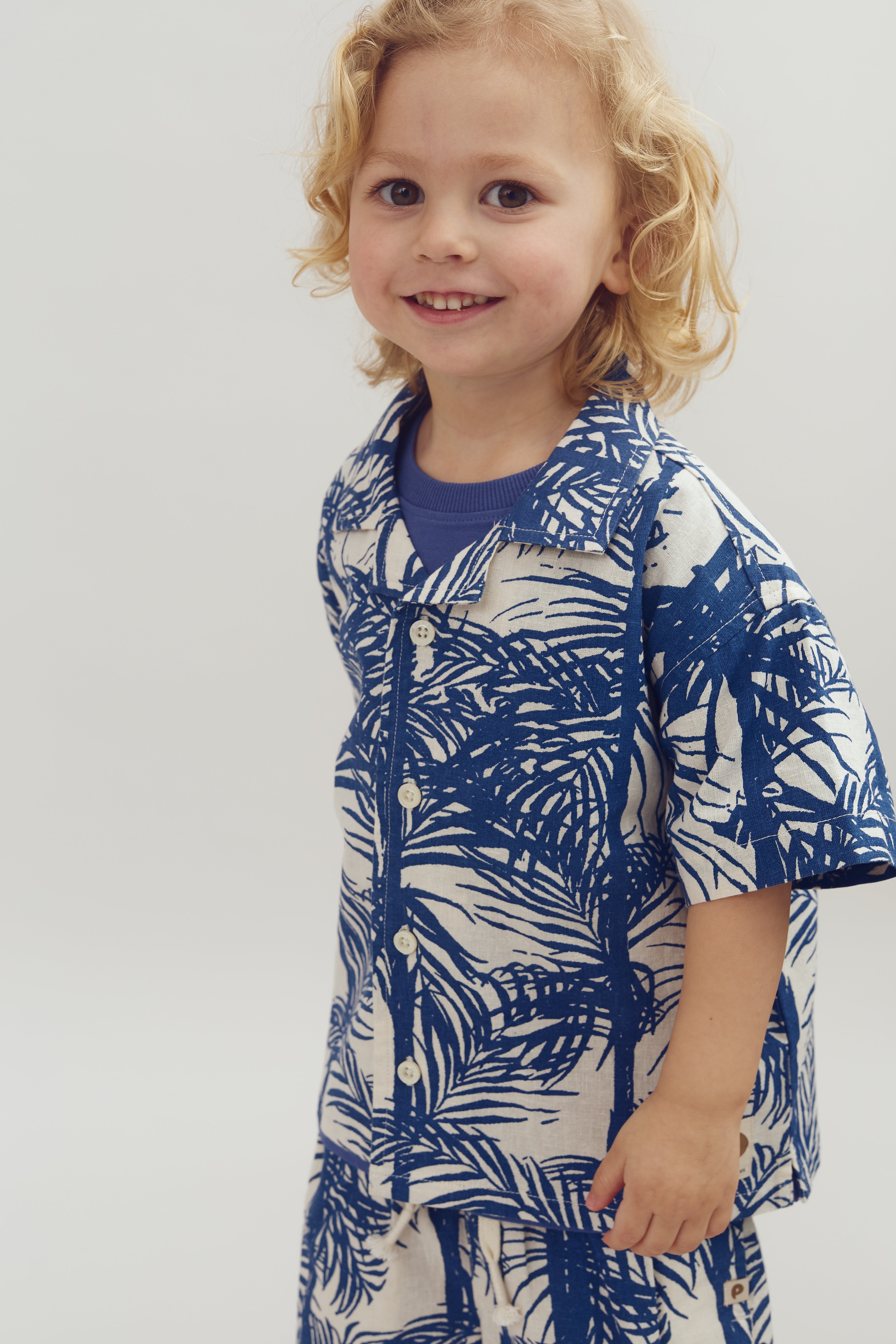 A young child with curly blond hair smiles, wearing a blue-and-white patterned shirt with palm designs, in a neutral, plain background.
