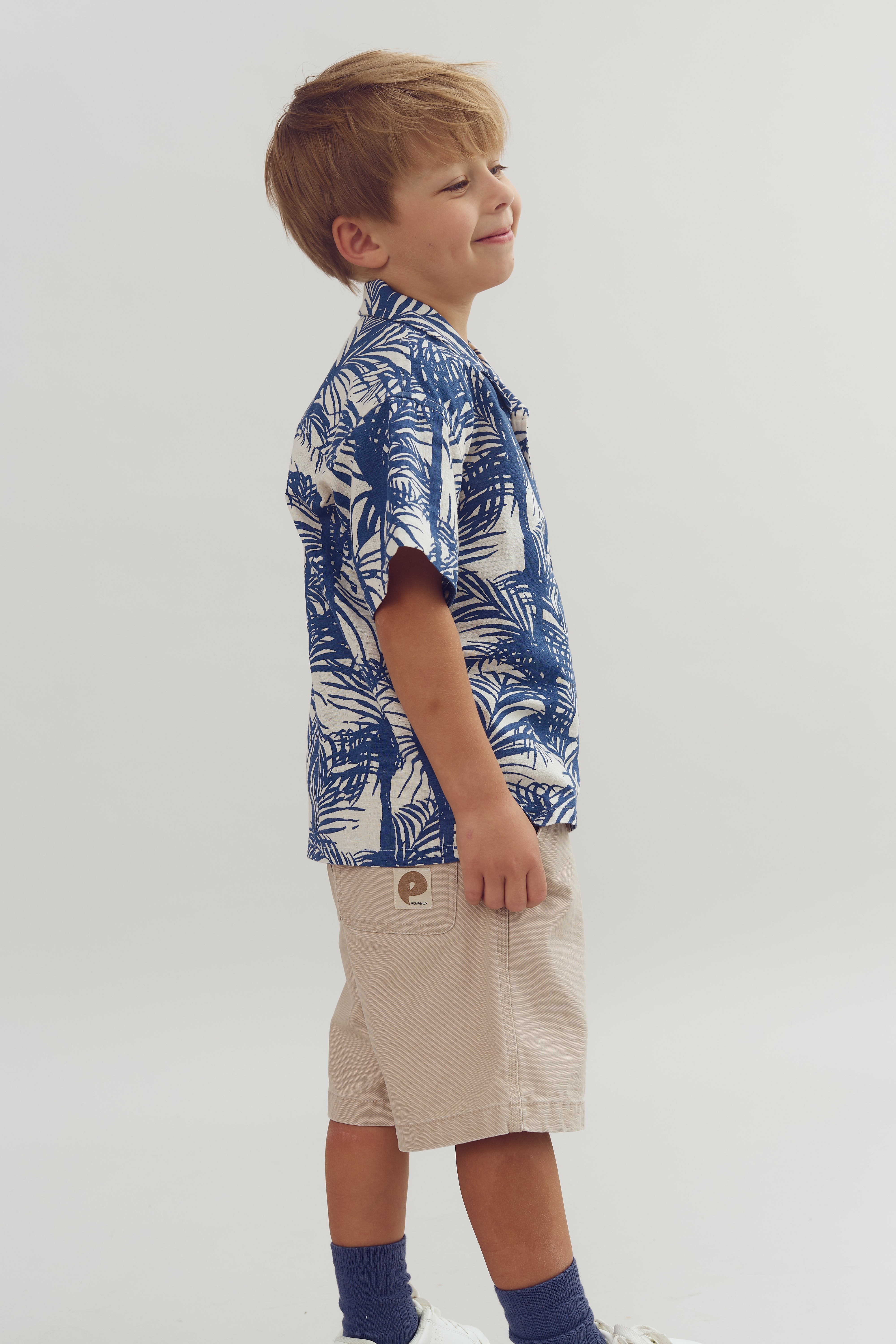 Dreng står smilende iført blå-hvid mønstret skjorte og beige shorts med et mærke på lommen; baggrund er neutral og hvid.