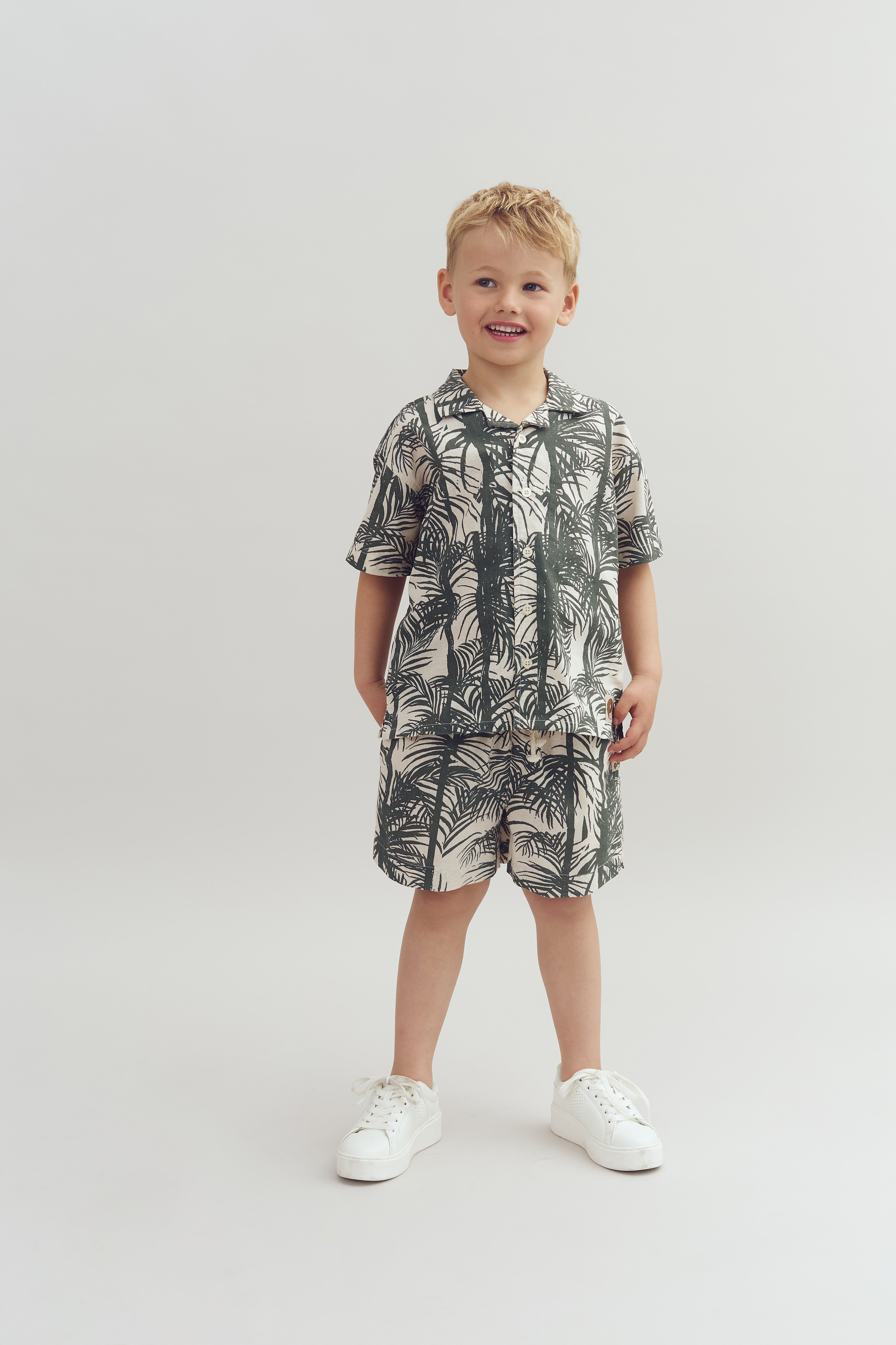A young child stands smiling, wearing a matching tropical-patterned shirt and shorts, with white sneakers. The background is a plain, light-colored studio setting.