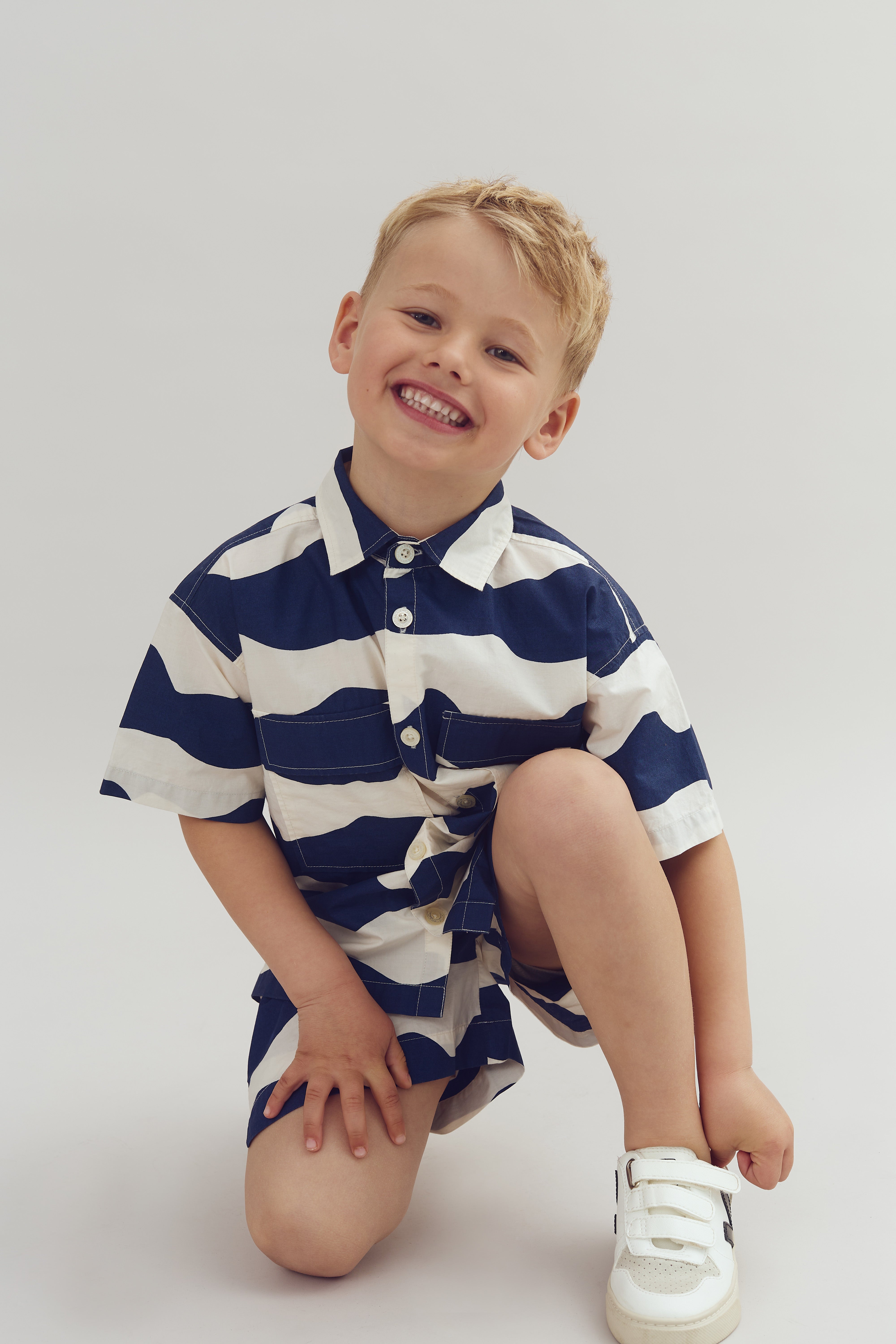 A child kneels, smiling widely, in a striped navy and white outfit and white velcro sneakers against a plain background.