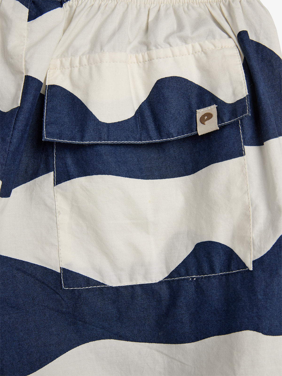 A garment featuring a pocket with a buttoned flap, displaying a navy and white wavy striped pattern. A small tag with a circular design is attached to the pocket flap.