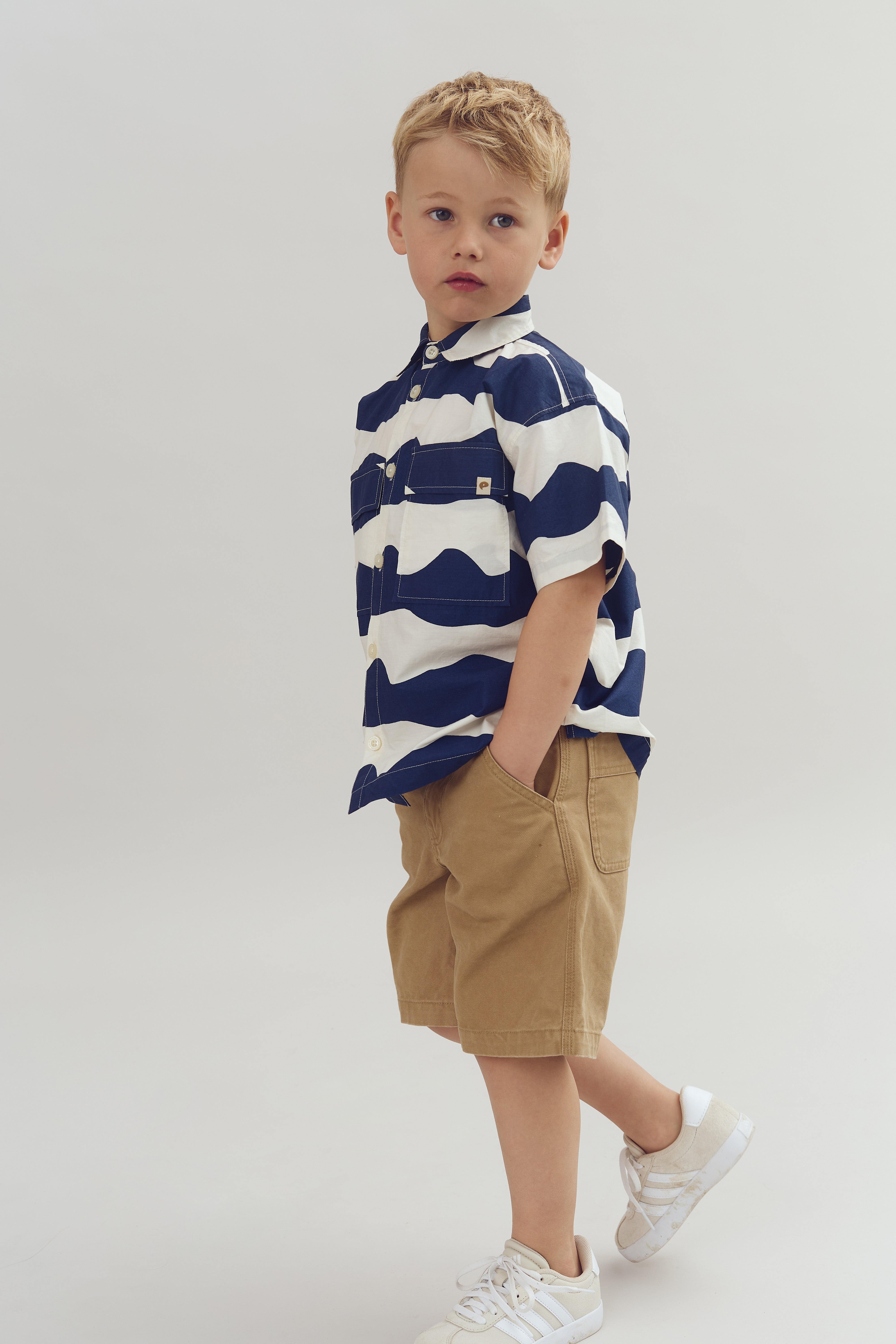 A young child stands with one hand in pocket, wearing a striped shirt, tan shorts, and white sneakers in a neutral studio setting.