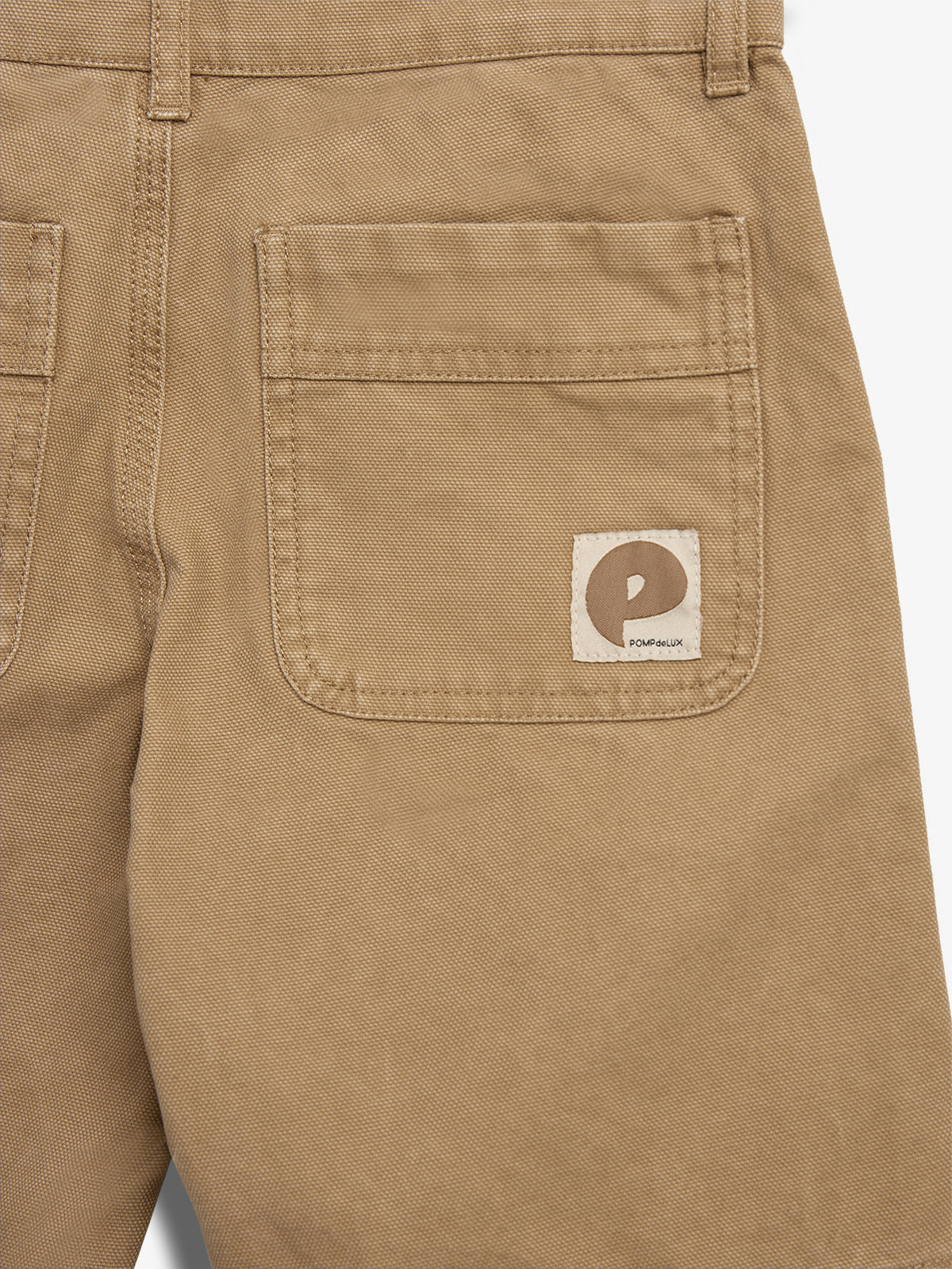 Khaki pants display a rear pocket with a sewn-on label featuring a stylized "P" and the text "POMPdeLUX" beneath. The background is plain and neutral-colored.