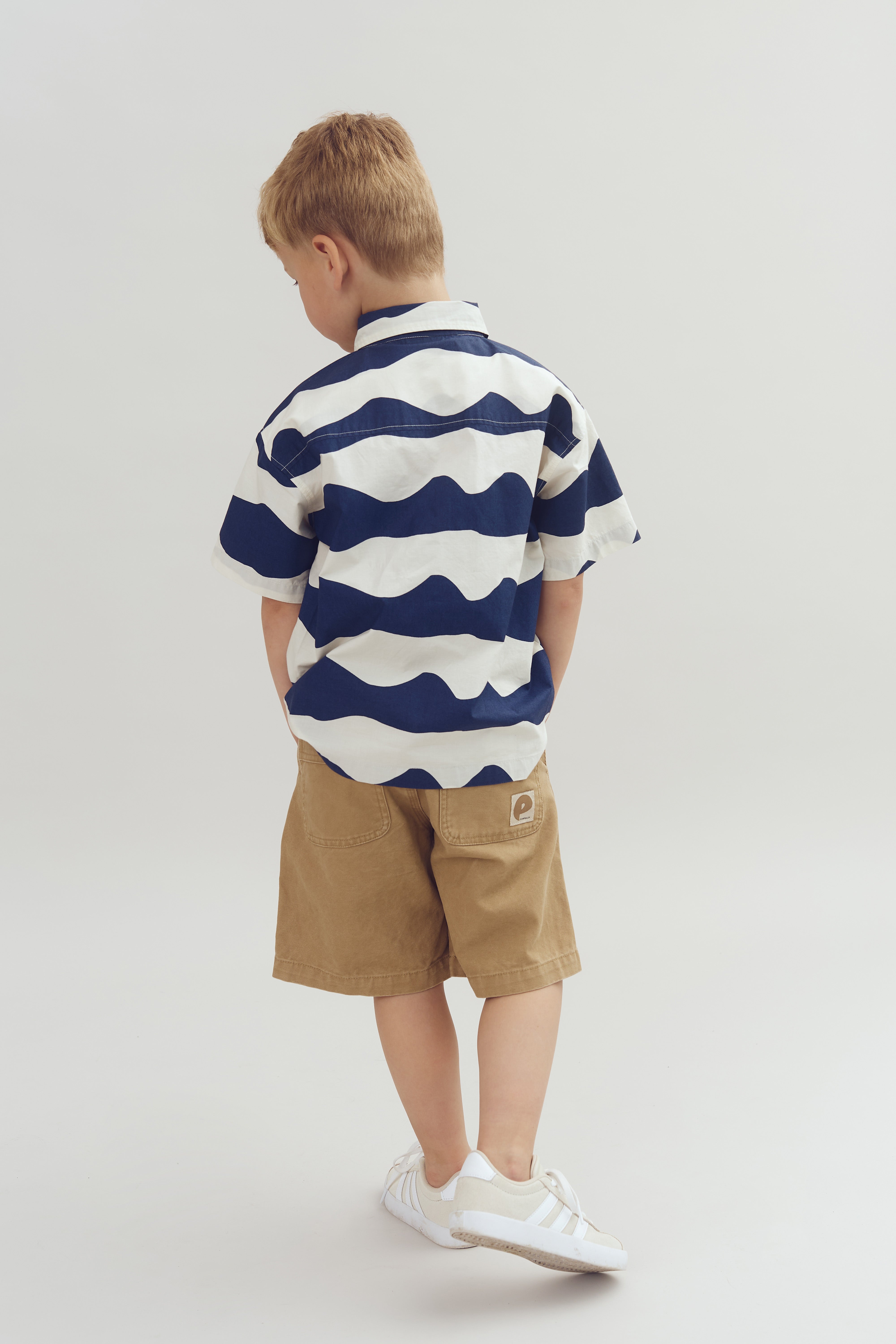 A child stands facing away, wearing a navy and white striped shirt, khaki shorts with a logo patch, and white sneakers. The background is a plain light gray.