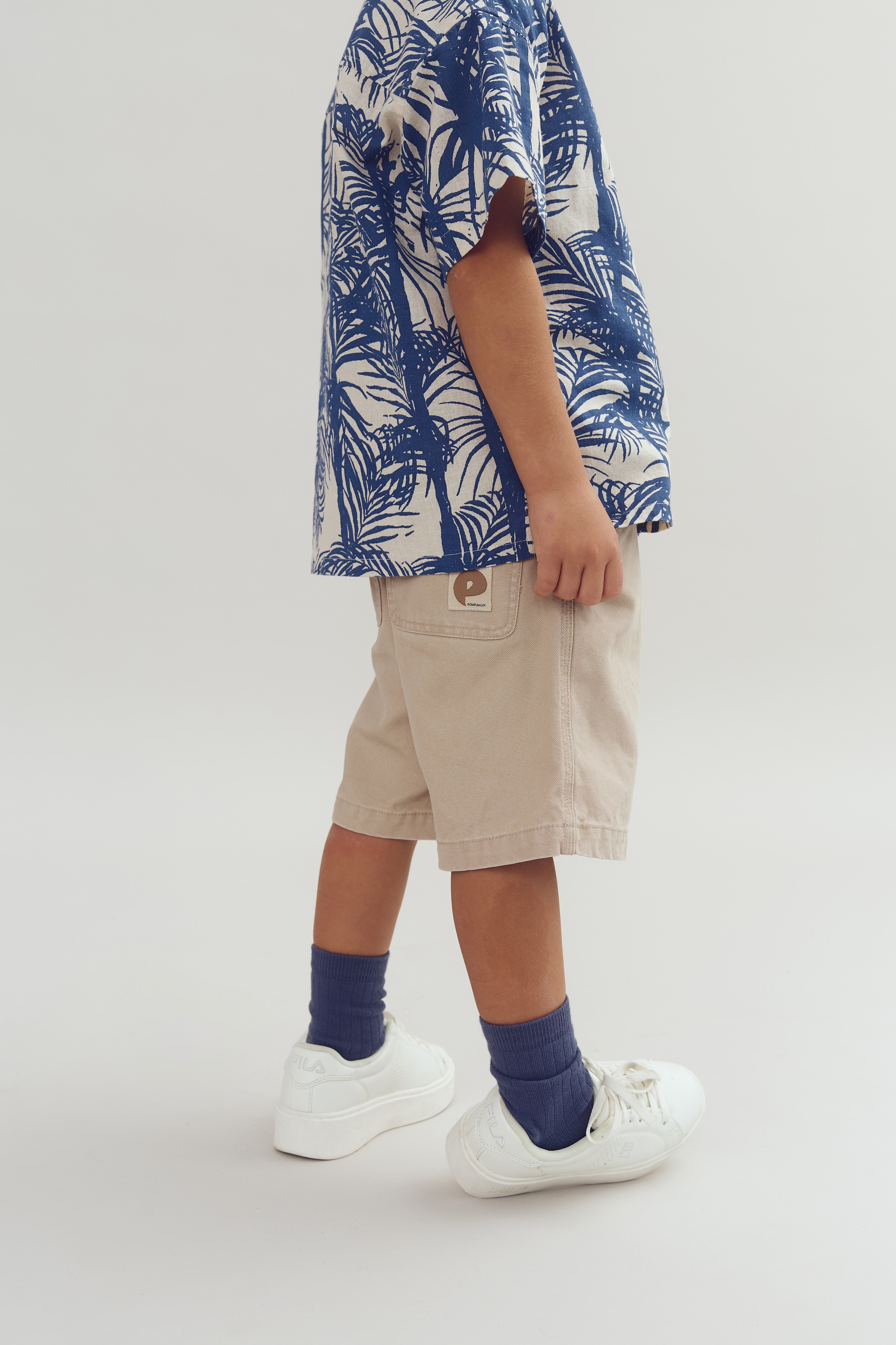 Barn står iført beige shorts og blå-hvid mønstret skjorte, med hvide sko og blå sokker, mod en neutral baggrund. Shortsene har et mærke med teksten "p".