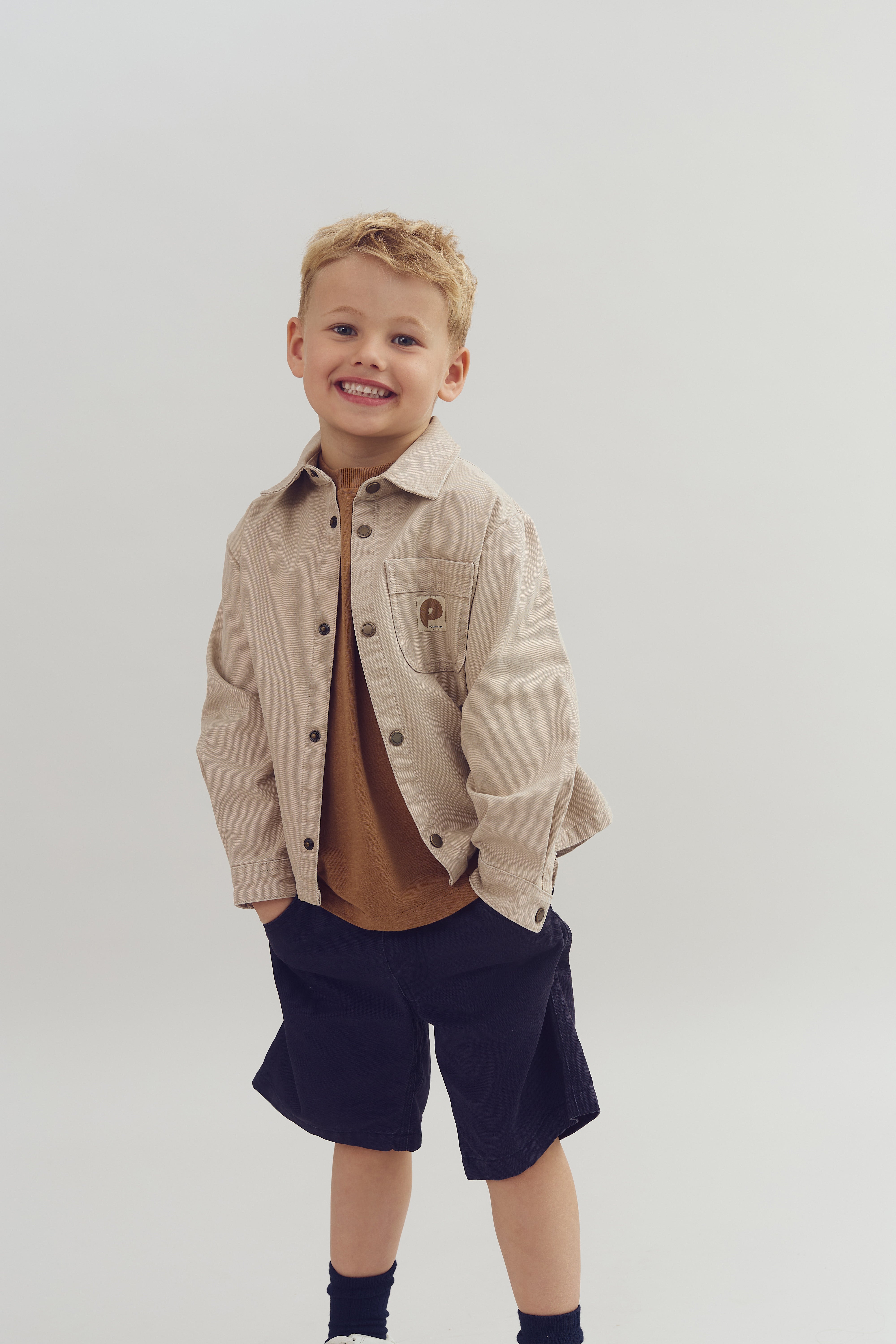 Dreng smiler iført beige jakke og sorte shorts. Han står med hænderne i lommerne mod en neutral baggrund.