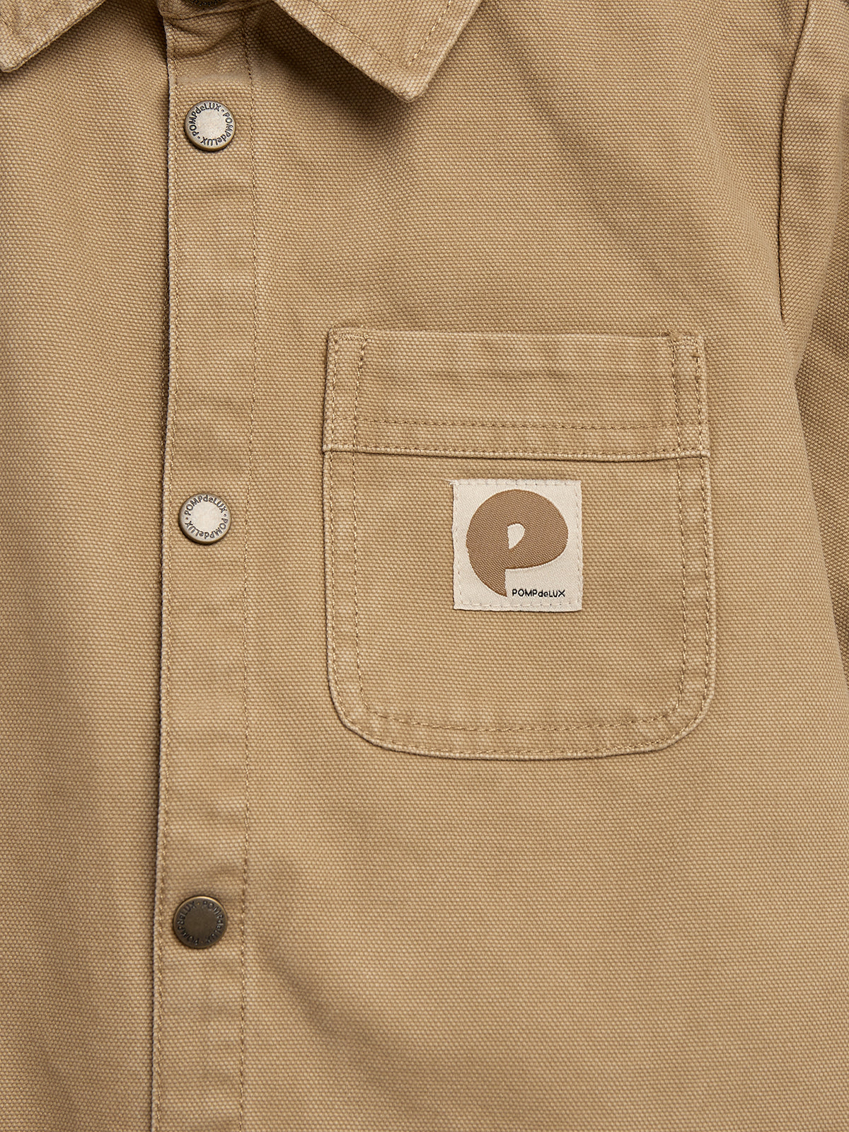 A beige shirt features metal snap buttons and a pocket on the chest. The pocket displays a small tag with a stylized "P" and text "POMPdeLUx" beneath it.