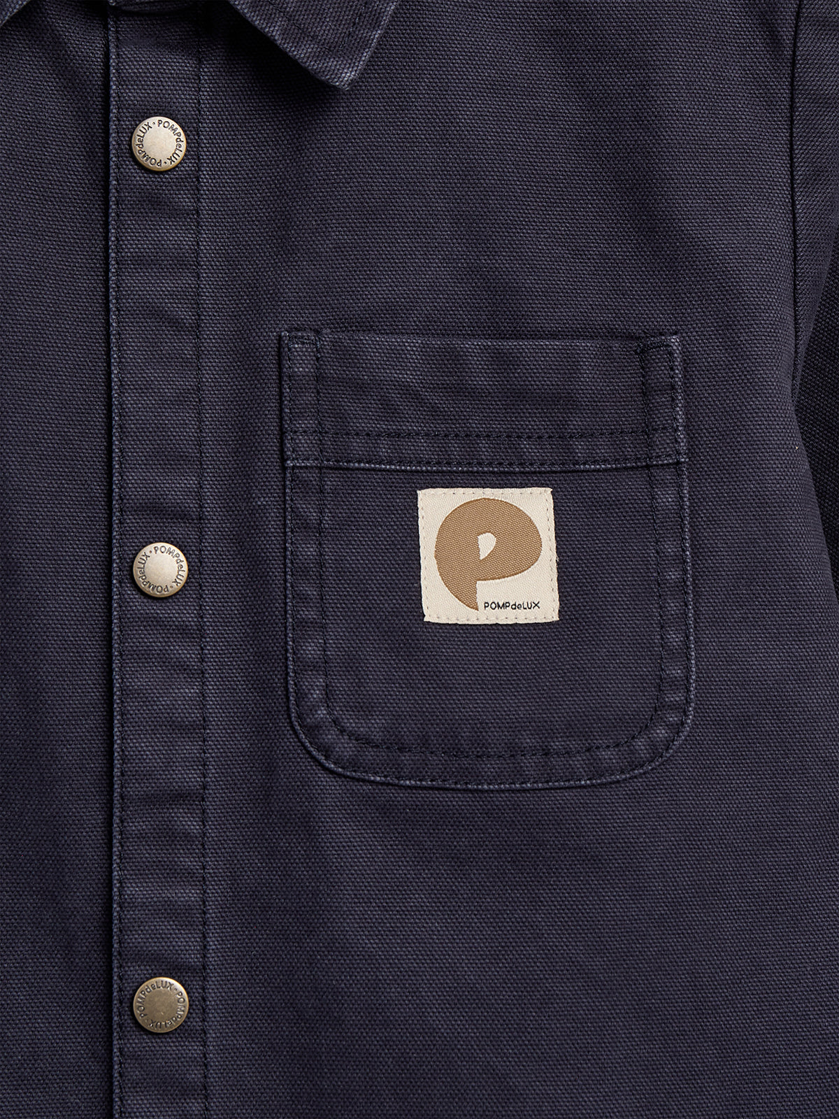 Dark blue shirt with silver buttons runs vertically down the front. A patch pocket on the left side features a beige square label with a brown "P" and text "POMPdeLUX."
