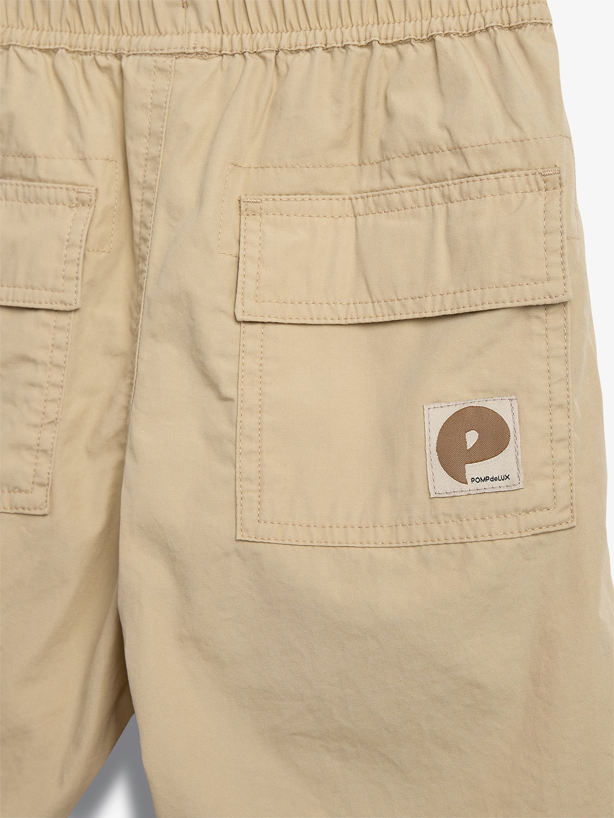 Beige shorts med to baglommer, hvoraf den ene har et logo med et bogstav "P". Teksten på logoet siger "POPMEDLUX". Shortsene er placeret mod en lys baggrund.