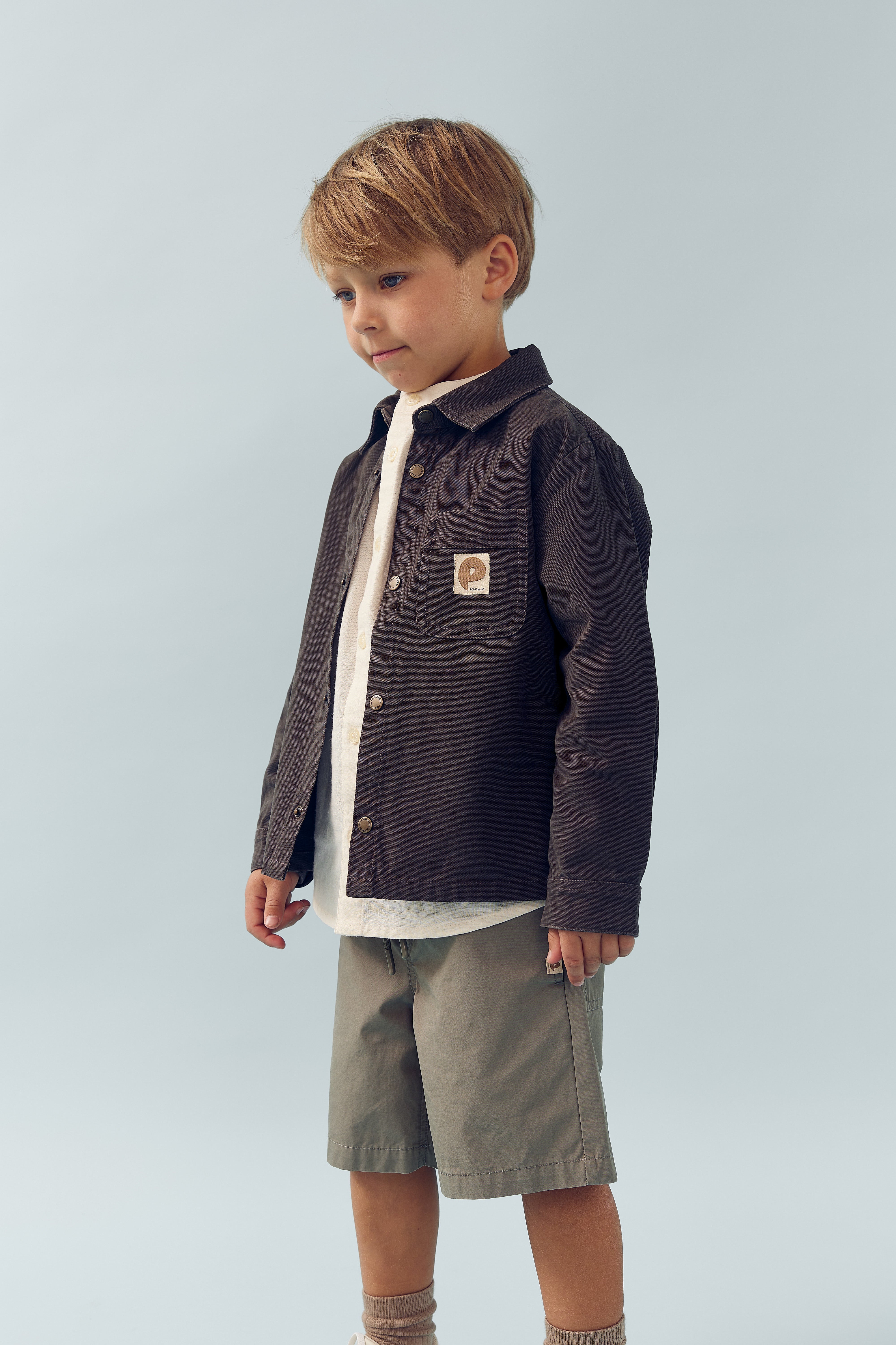 Et barn står stille iført en mørk jakke med en logo på brystlommen, lyse shorts og beige sokker. Baggrunden er lyseblå.