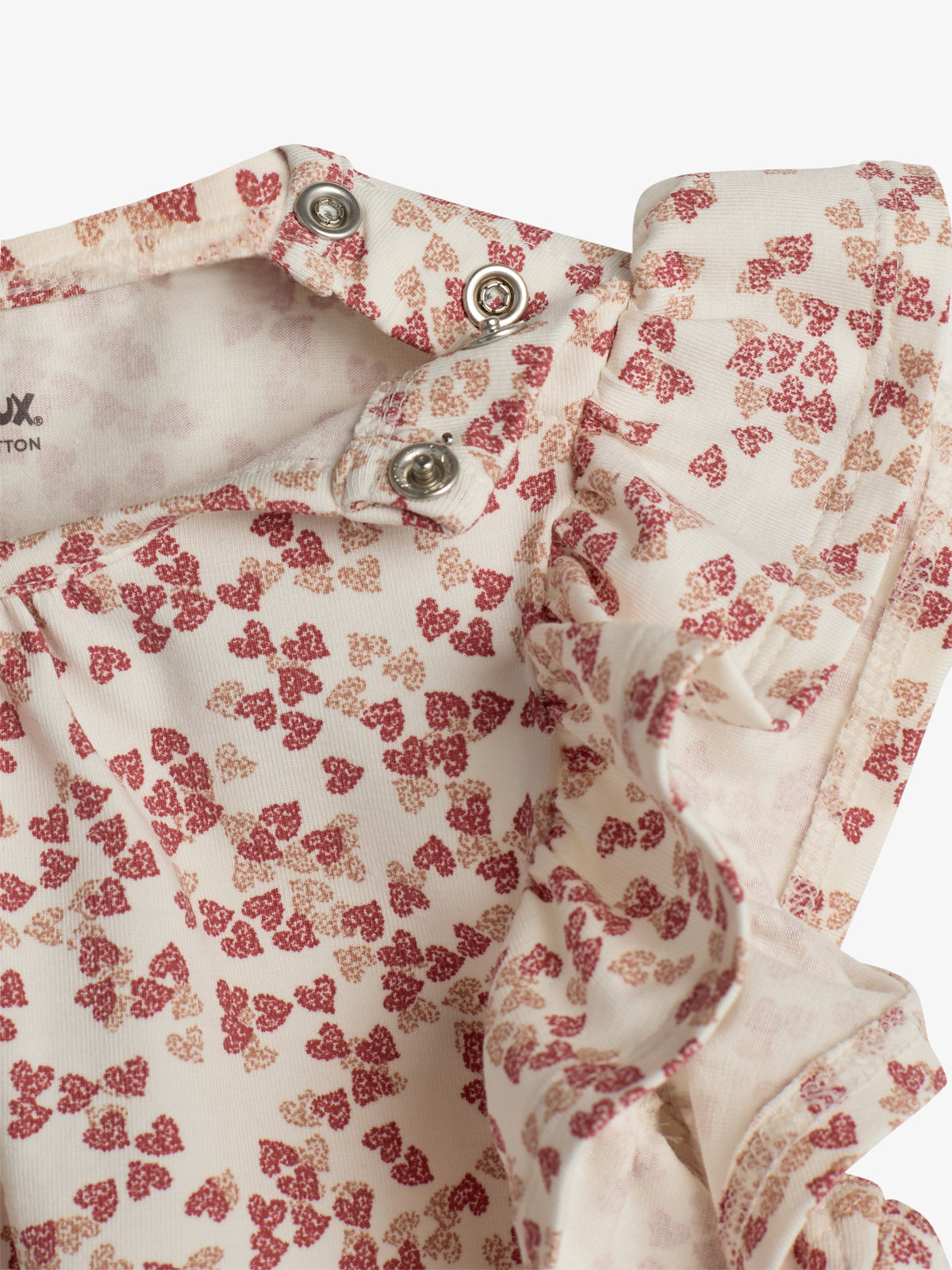 En hvid stofdel med røde og beige blomsterlignende mønstre, har metaltrykknapper ved åbningen. Stoffet er let rynket, hvilket tilføjer tekstur og bevægelse. Tekst: "XION. COTTON".