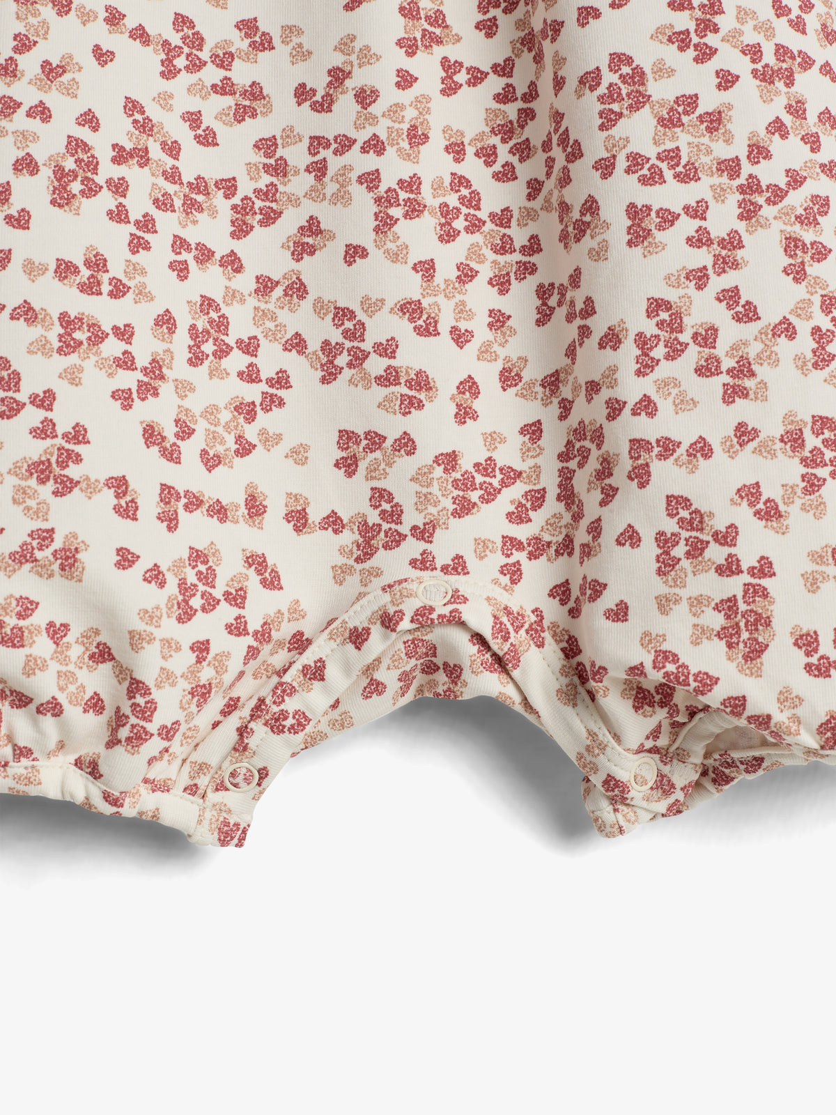 Et stykke tøj med små røde og lyserøde hjerteformede mønstre, viser en kant med trykknapper. Baggrunden er neutral og fremhæver stoffets design.