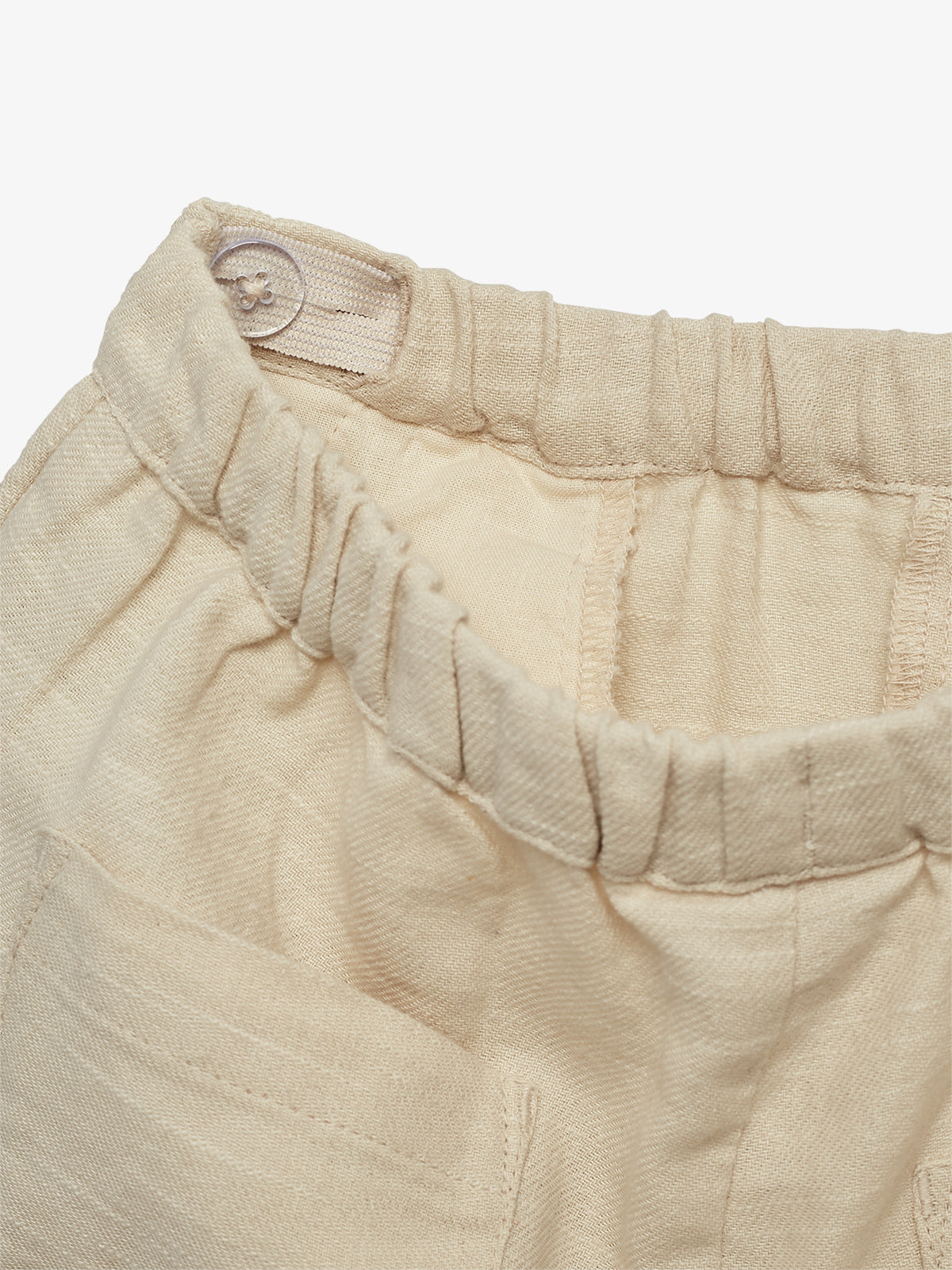Et par beige bukser med elastisk talje har en knapdetalje og synlige syninger, som fremhæves tæt på en neutral baggrund.