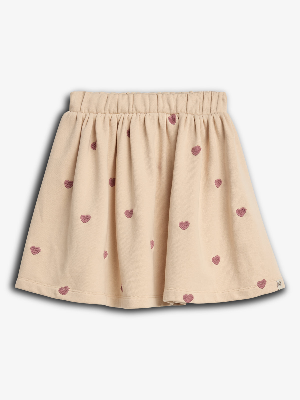 En beige nederdel med elastisk talje og små pink hjerteprint spredt over stoffet. Baggrunden er ensfarvet lys grå, hvilket fremhæver nederdelens detaljerede design.
