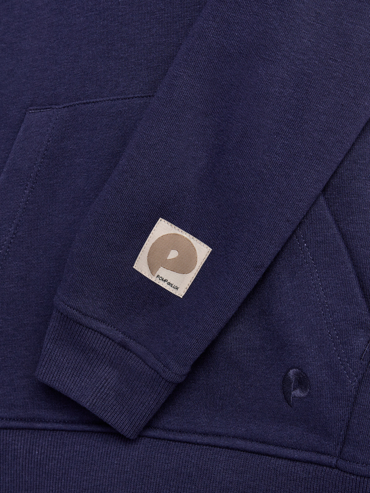 A navy blue sweatshirt sleeve with a beige patch featuring a circular design and the text "POMPdeLUX." The sleeve rests on the body of the sweatshirt, which has visible stitching.