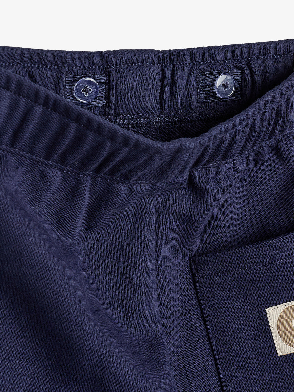 Navy blue elastic waistband with button closures is attached to a pair of pants. Nearby, a flat pocket is partially visible, featuring a beige rectangular label with a circular pattern.