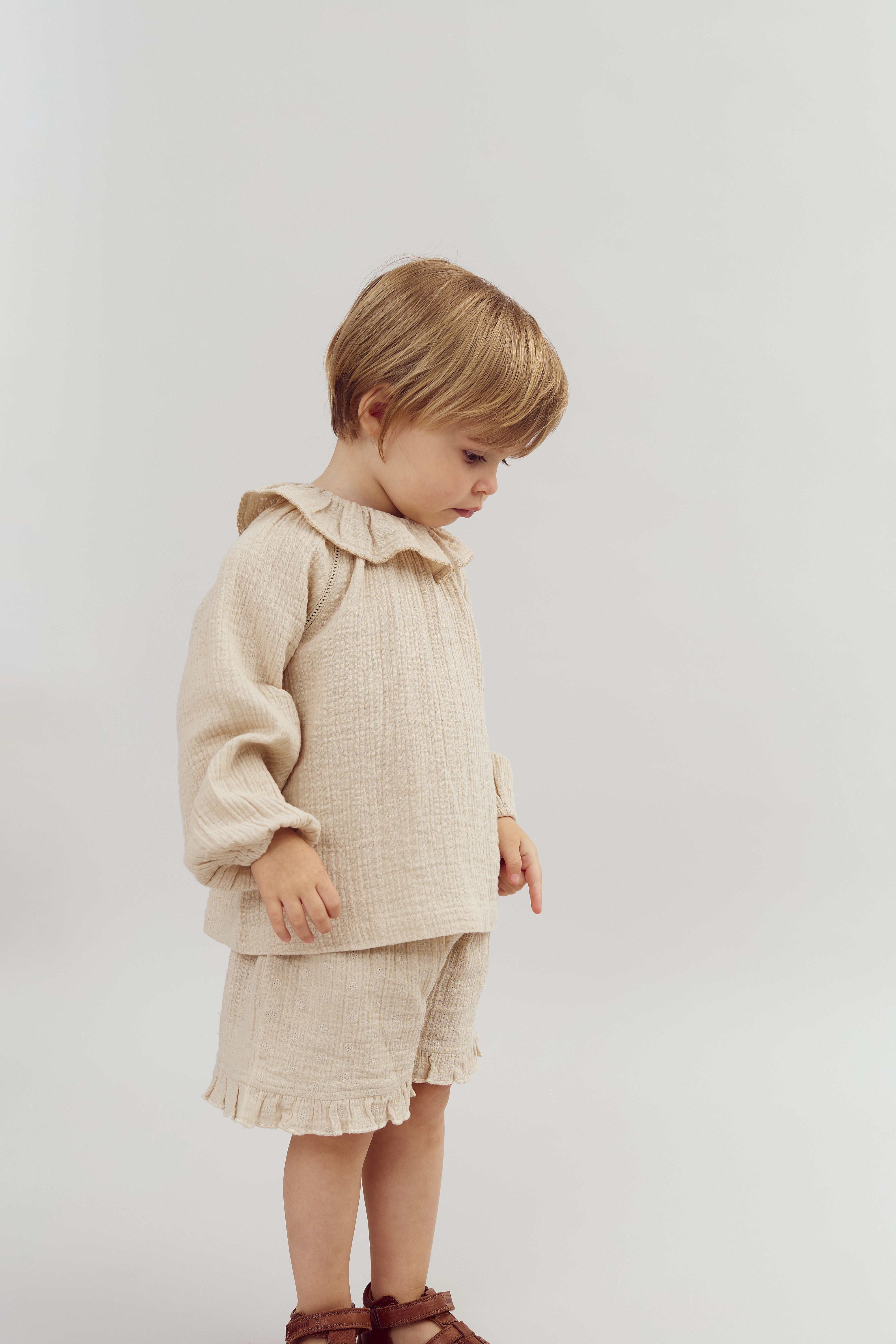 A child stands in a beige outfit with ruffled details, looking downwards in a neutral, light-colored environment.
