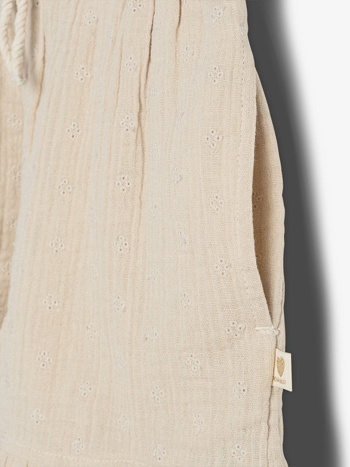 Beige fabric with small embroidered floral patterns features a side pocket. A small tag with a heart and "Mini Rodini" hangs near the seam, set against a neutral background.