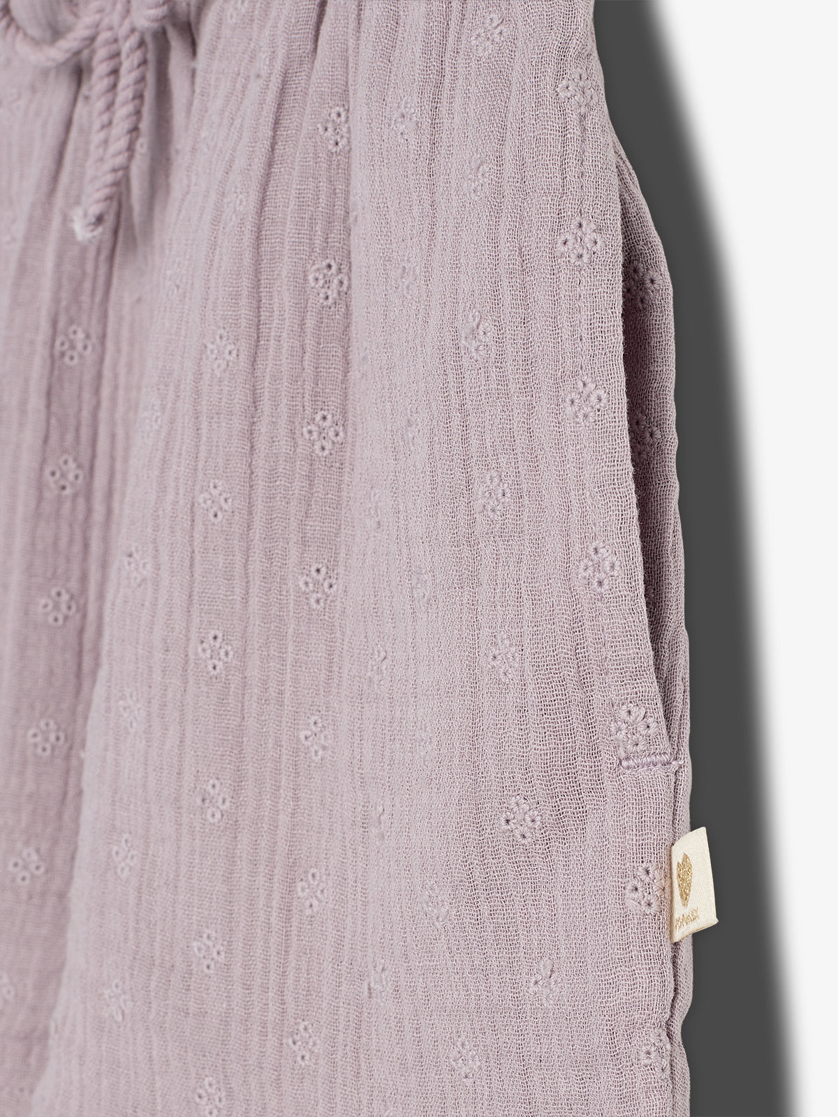 A pair of soft, textured lavender pants with a drawstring is shown hanging. The pants have a subtle floral pattern, with a beige tag displaying a small leaf logo on the side.