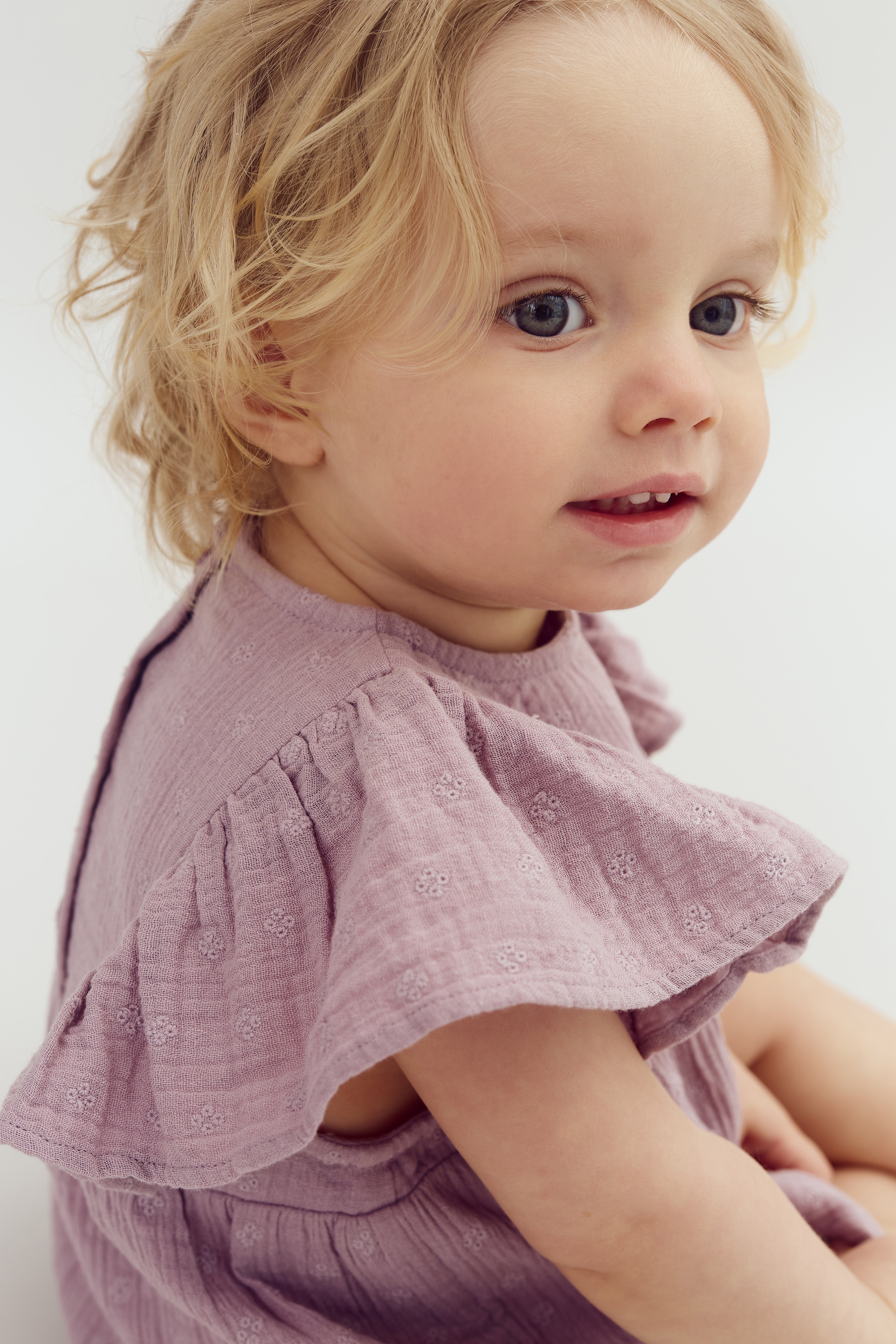 Toddler with light curly hair, looking to the side, wearing a lavender dress with embroidered patterns, sitting in a softly lit, neutral environment.
