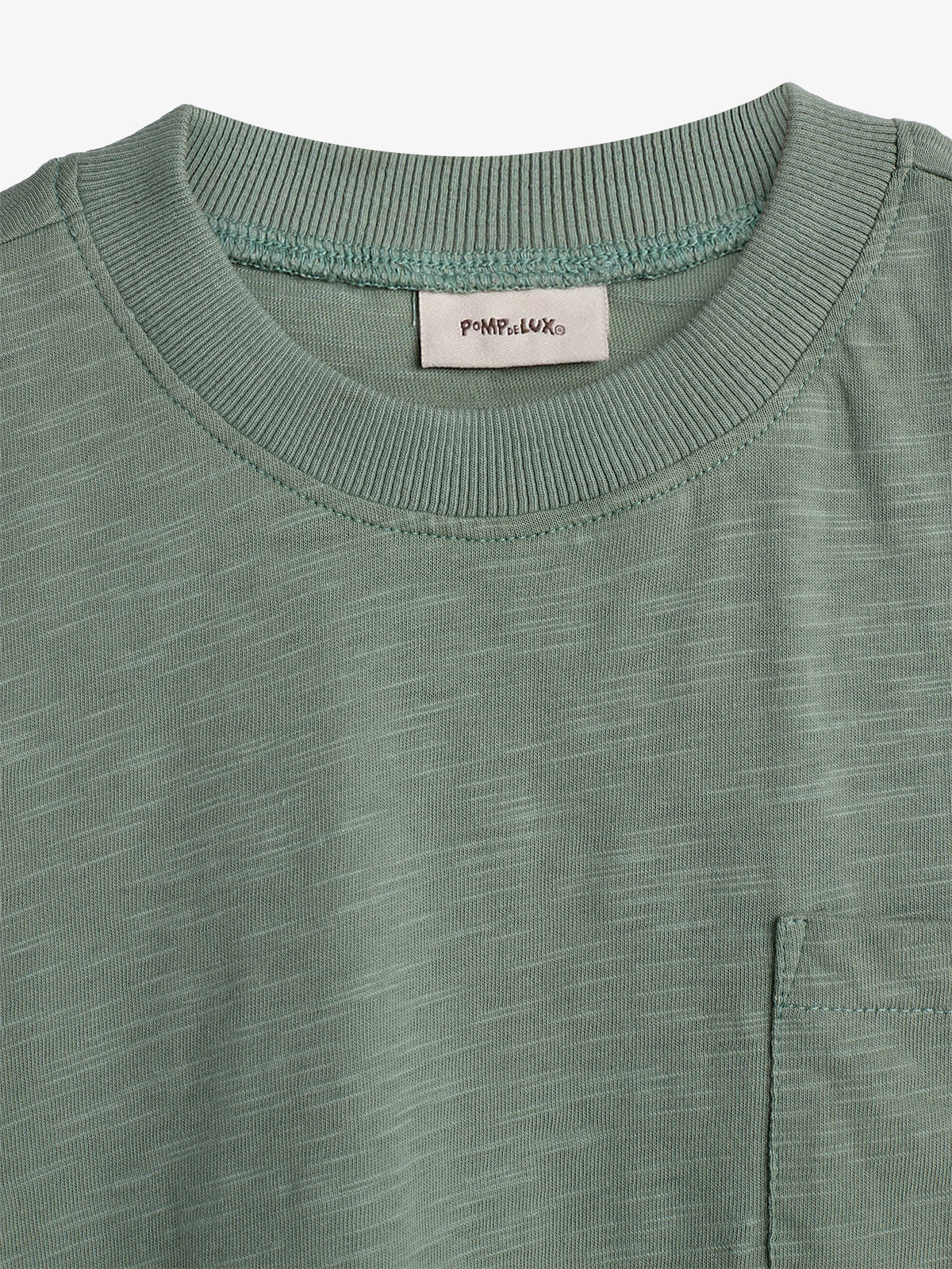 A green T-shirt with ribbed collar and stitched pocket sits against a plain background. Text on label reads "POMPdeLUX."