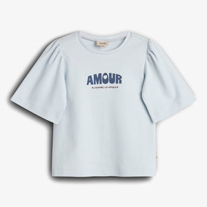 A short-sleeved, light blue shirt with puffed sleeves displays the text "AMOUR" in bold, large letters and "J'ADORE LE SOLEIL" beneath. The shirt is on a plain background.
