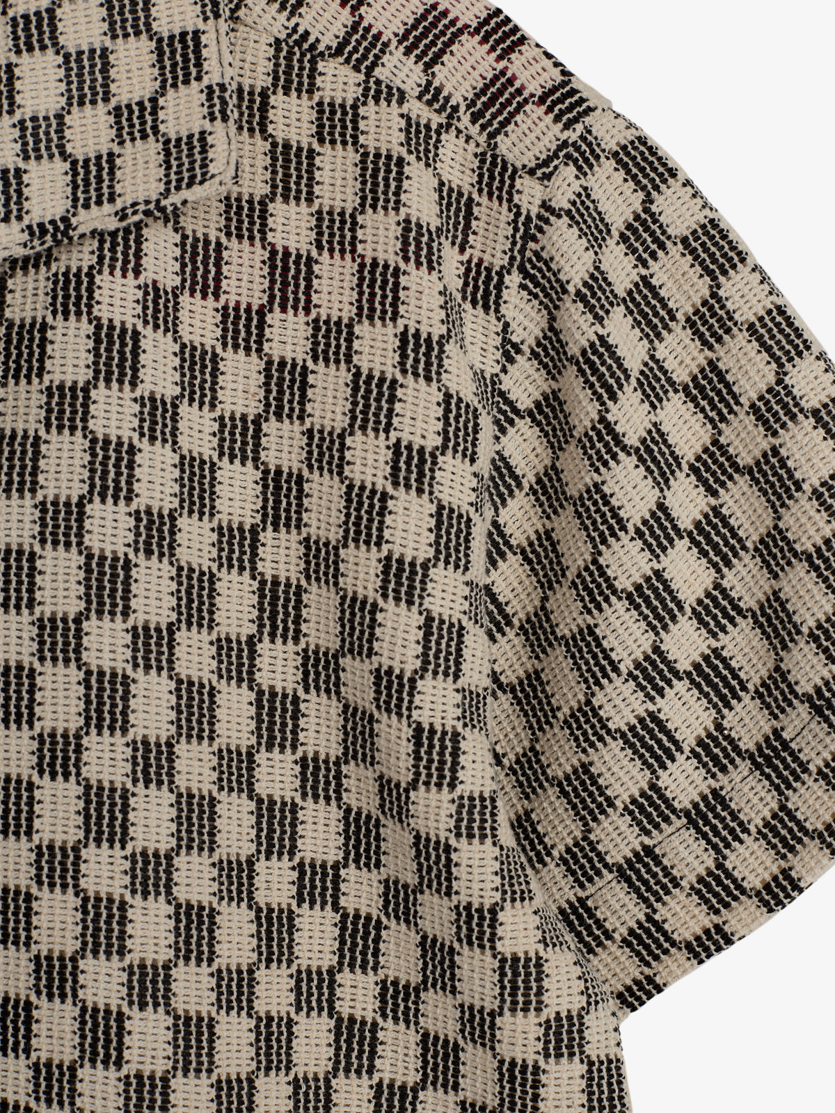 Skjorte med sort-hvid ternet mønster vises i tæt detalje. Den har en krave og korte ærmer, og stoffet fremstår vævet. Baggrunden er ensfarvet lys.