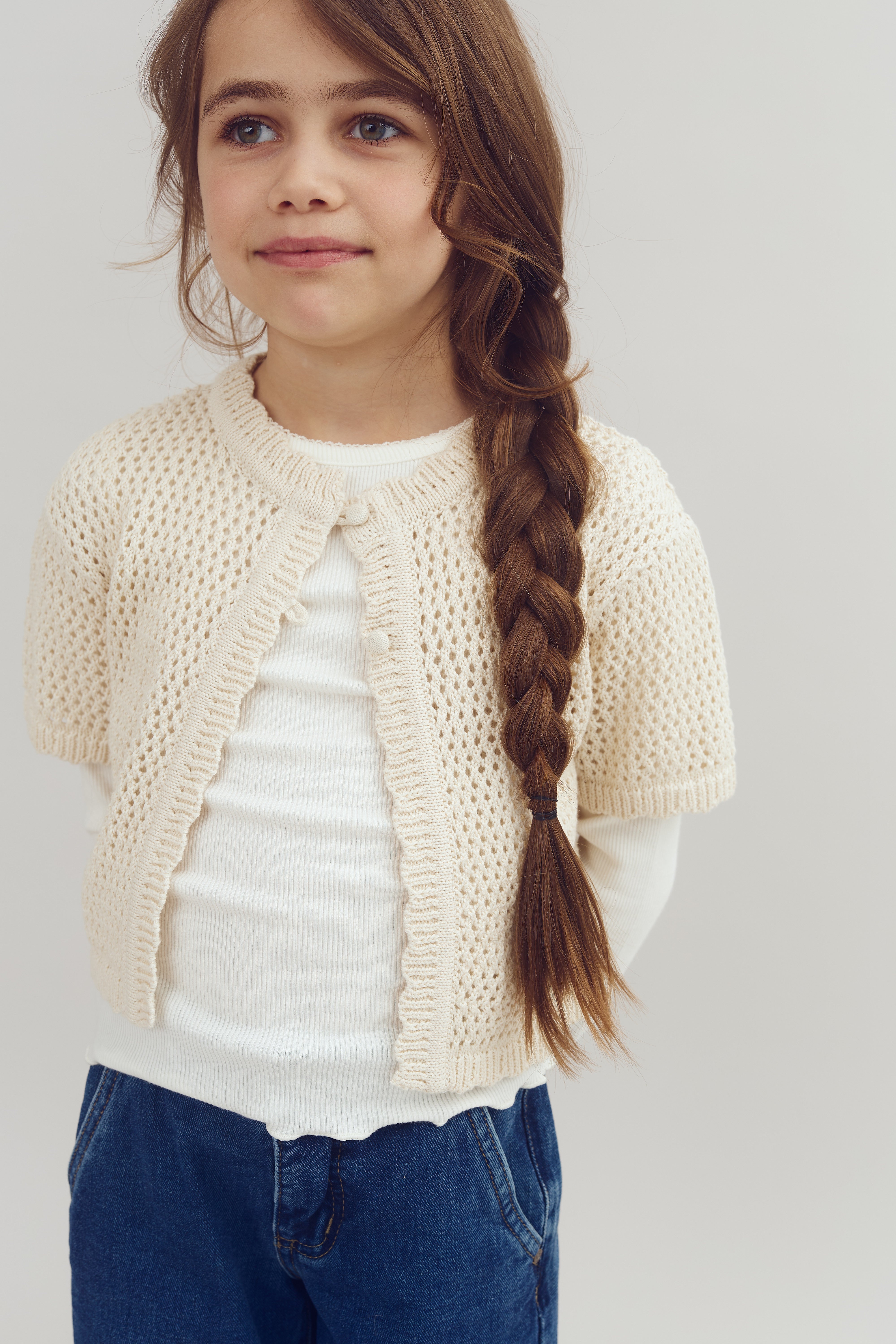A young girl stands smiling, wearing a white knit cardigan, white shirt, and blue jeans, her long brown hair in a braid, against a plain light background.