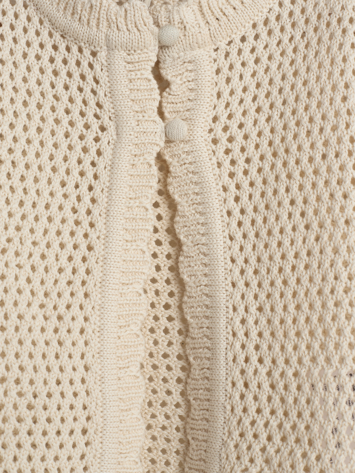 A cream-colored knitted cardigan features a textured, open-weave pattern with two small round buttons along a ribbed edge, creating a detailed, delicate appearance.
