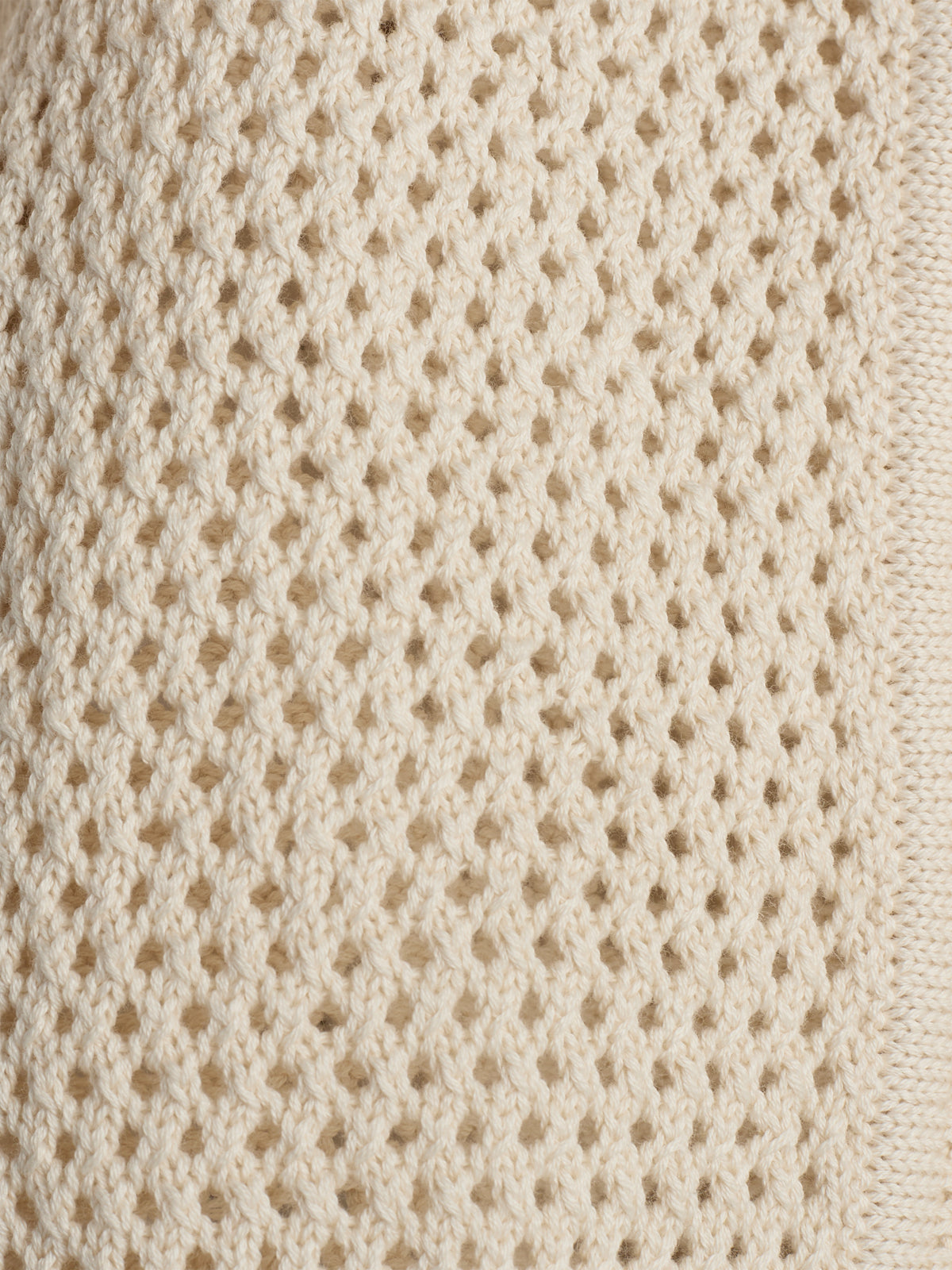 Beige knitted fabric with a honeycomb pattern shows an intricate, uniform texture, creating small diamond-shaped holes, providing a cozy and delicate appearance. The fabric is lit in soft natural light.