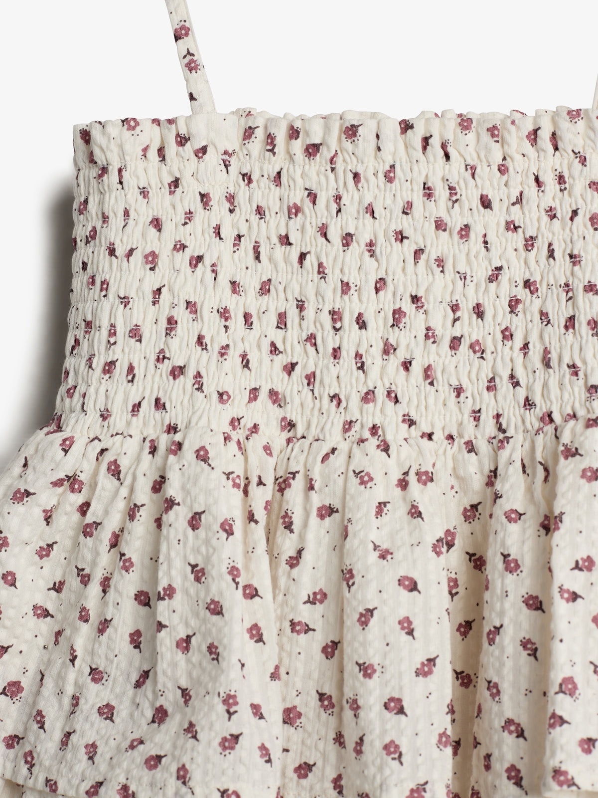 A smocked cream top, adorned with small red floral patterns, features thin straps. The fabric gathers above a loosely flowing section, set against a plain white background.