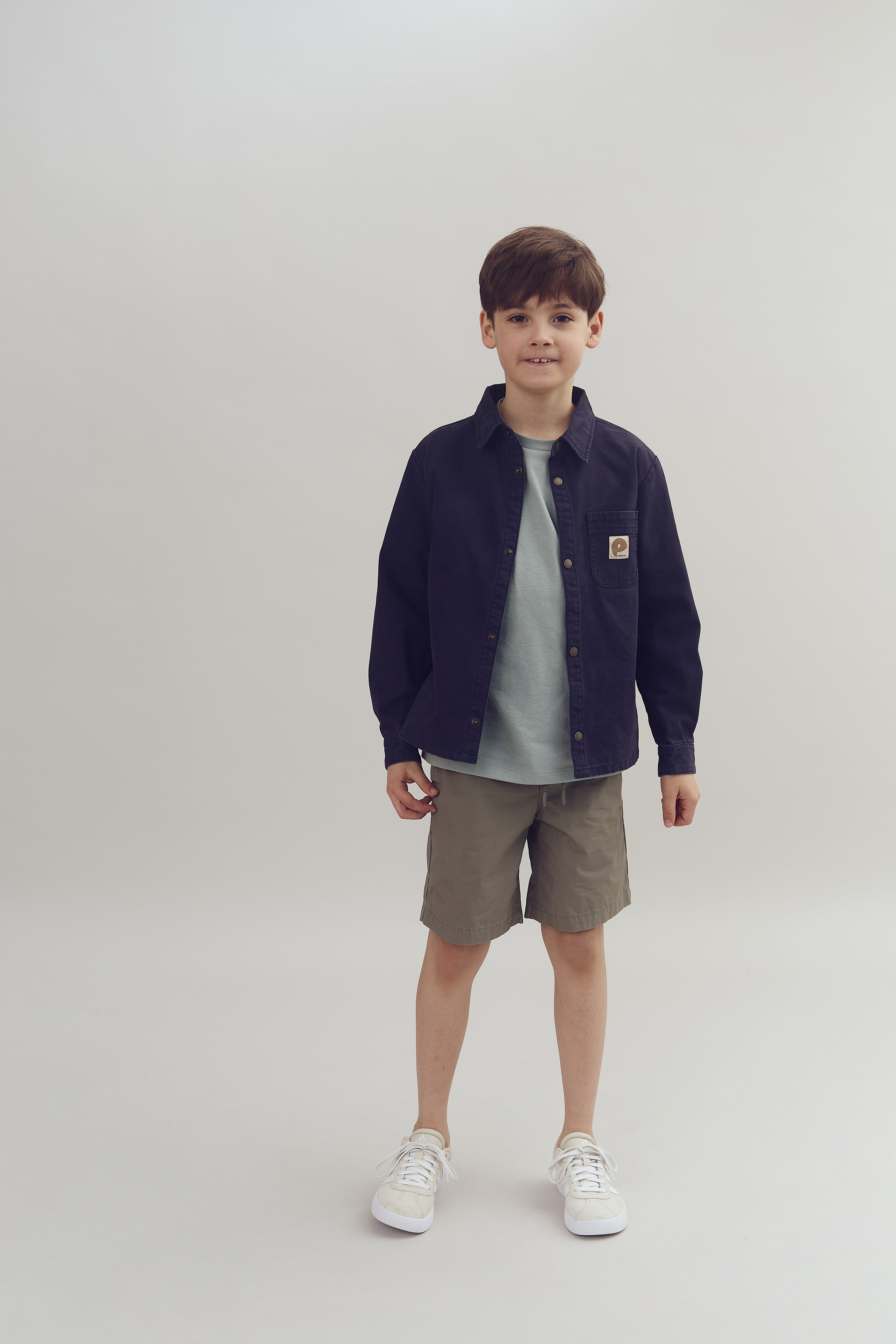 Dreng står iført mørk jakke, grå t-shirt, korte beige bukser og hvide sneakers. Han står i et neutralt, lyst studie.