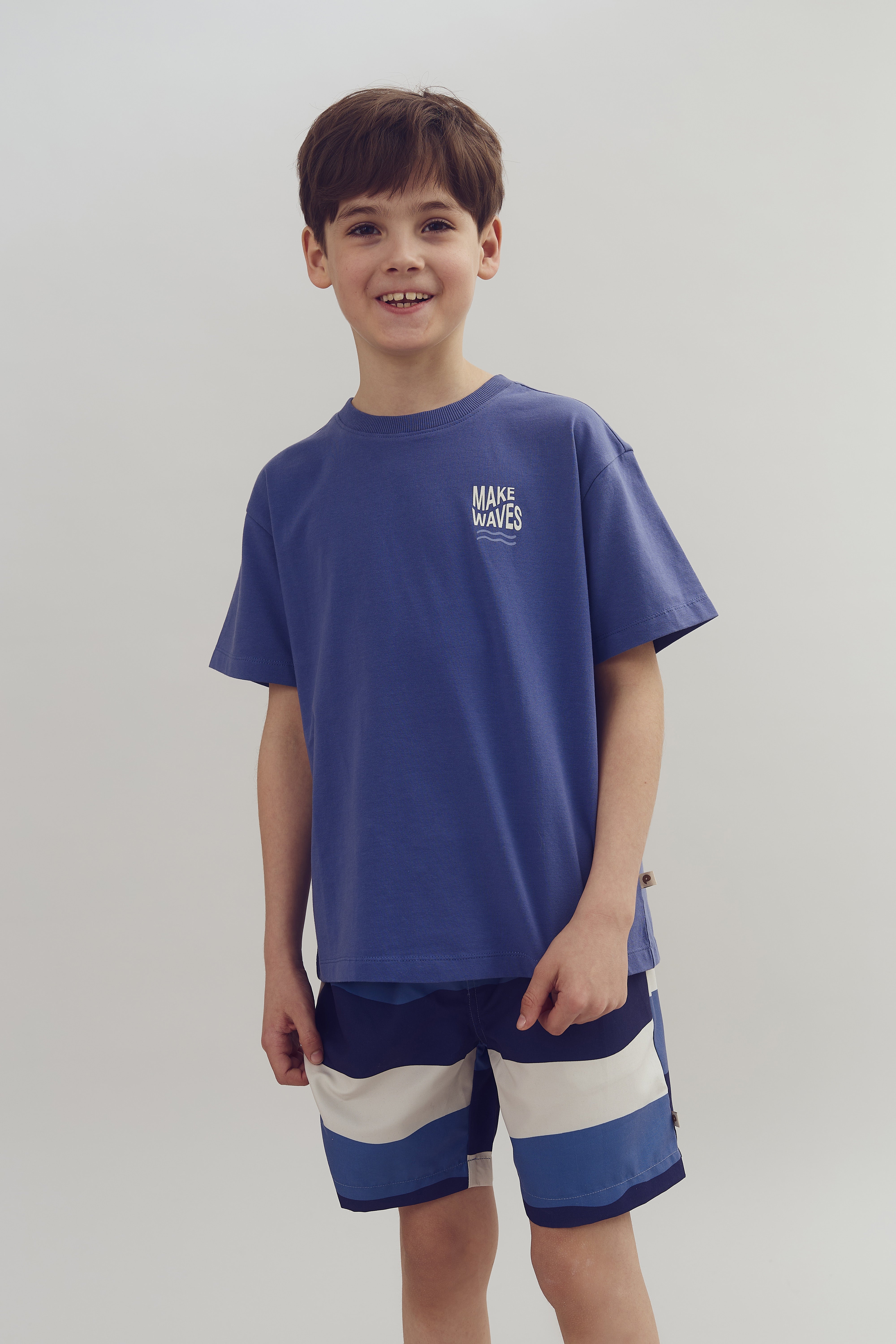 Dreng iført blå T-shirt med teksten "MAKE WAVES" og stribede shorts, står smilende i et neutralt rum.