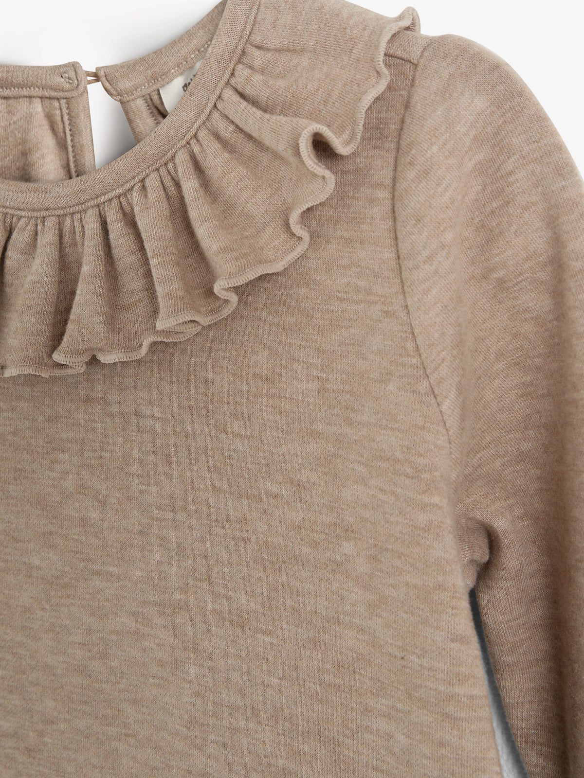 Beige trøje med flæset krave, hænger stille mod en hvid baggrund. Blød tekstur, lange ærmer, lukkes med lille knap i nakken. Trøjen er Enkel i designet, uden mønster.
