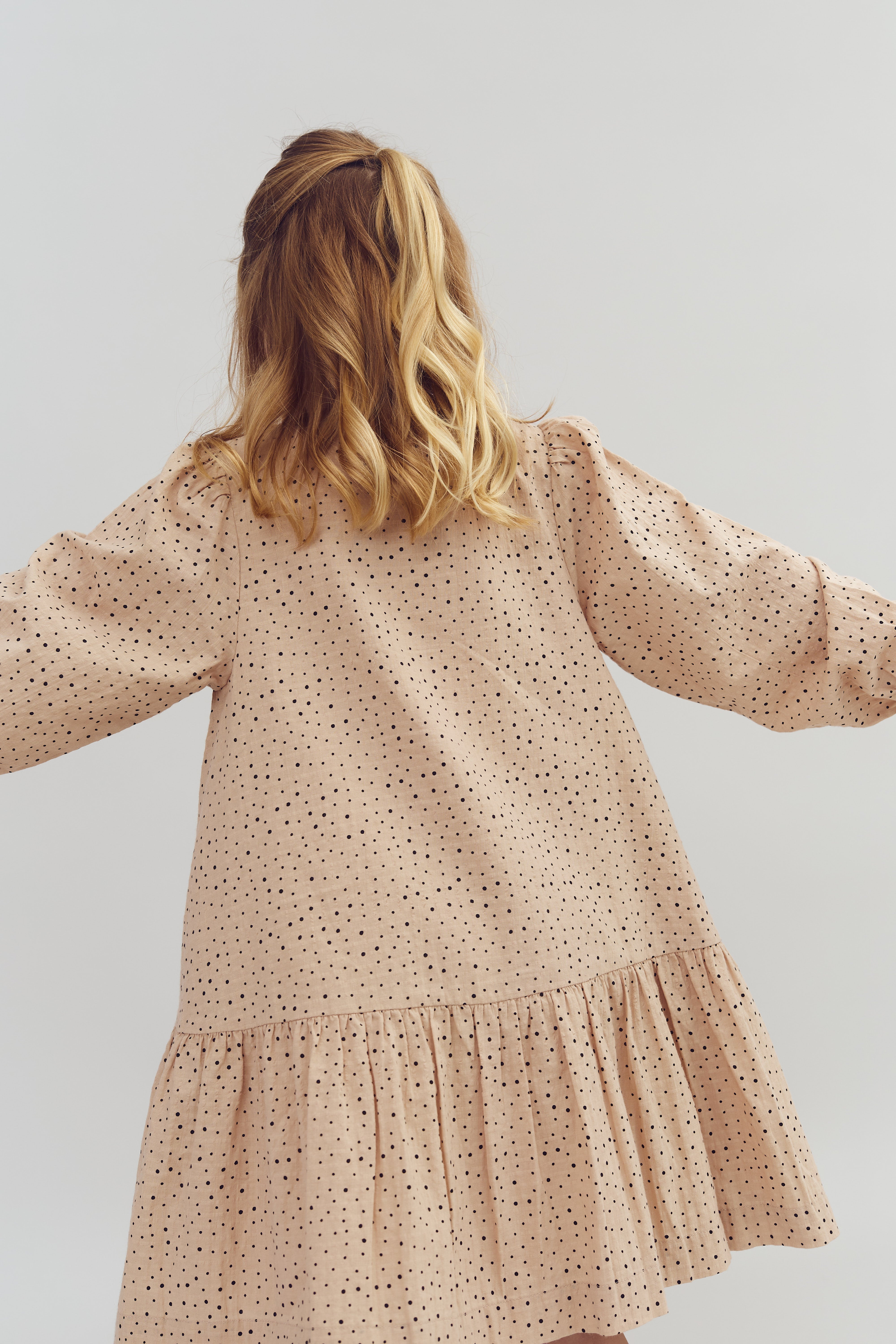 A person facing away wears a beige dress with black polka dots, standing against a plain, light background. Their hair is wavy and styled with a half-updo.