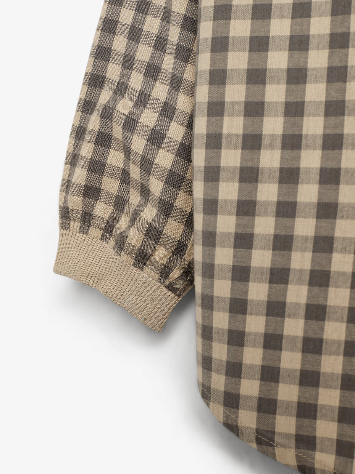 Ærmet på en ternet skjorte, med beige og mørkebrun farve, afsluttes med en ribstrikket manchet. Baggrunden er en ensfarvet, lysegrå overflade.