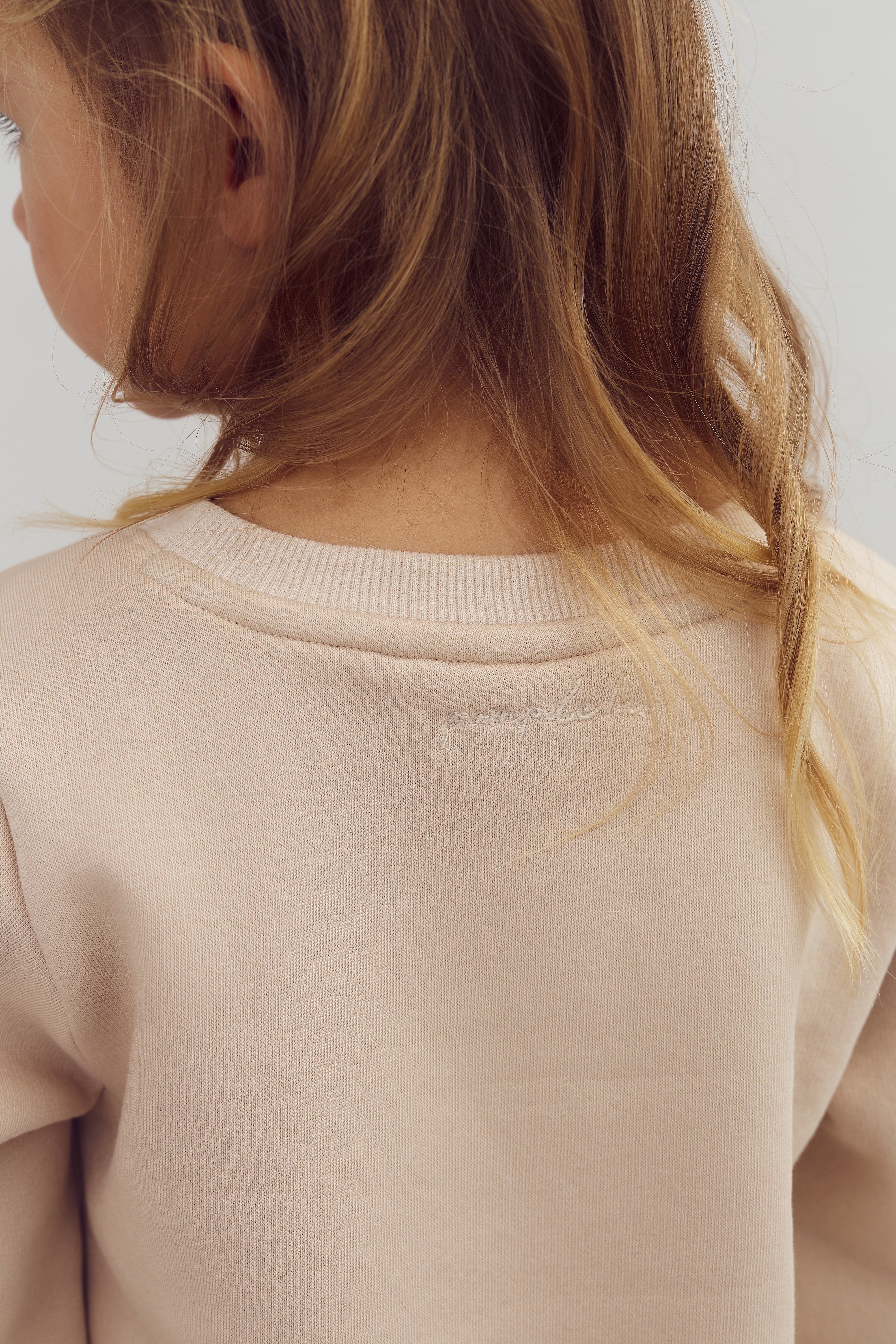 Barn iført beige trøje vendt væk, med langt, bølget lysebrunt hår; neutral baggrund. Subtil broderi på trøjen.