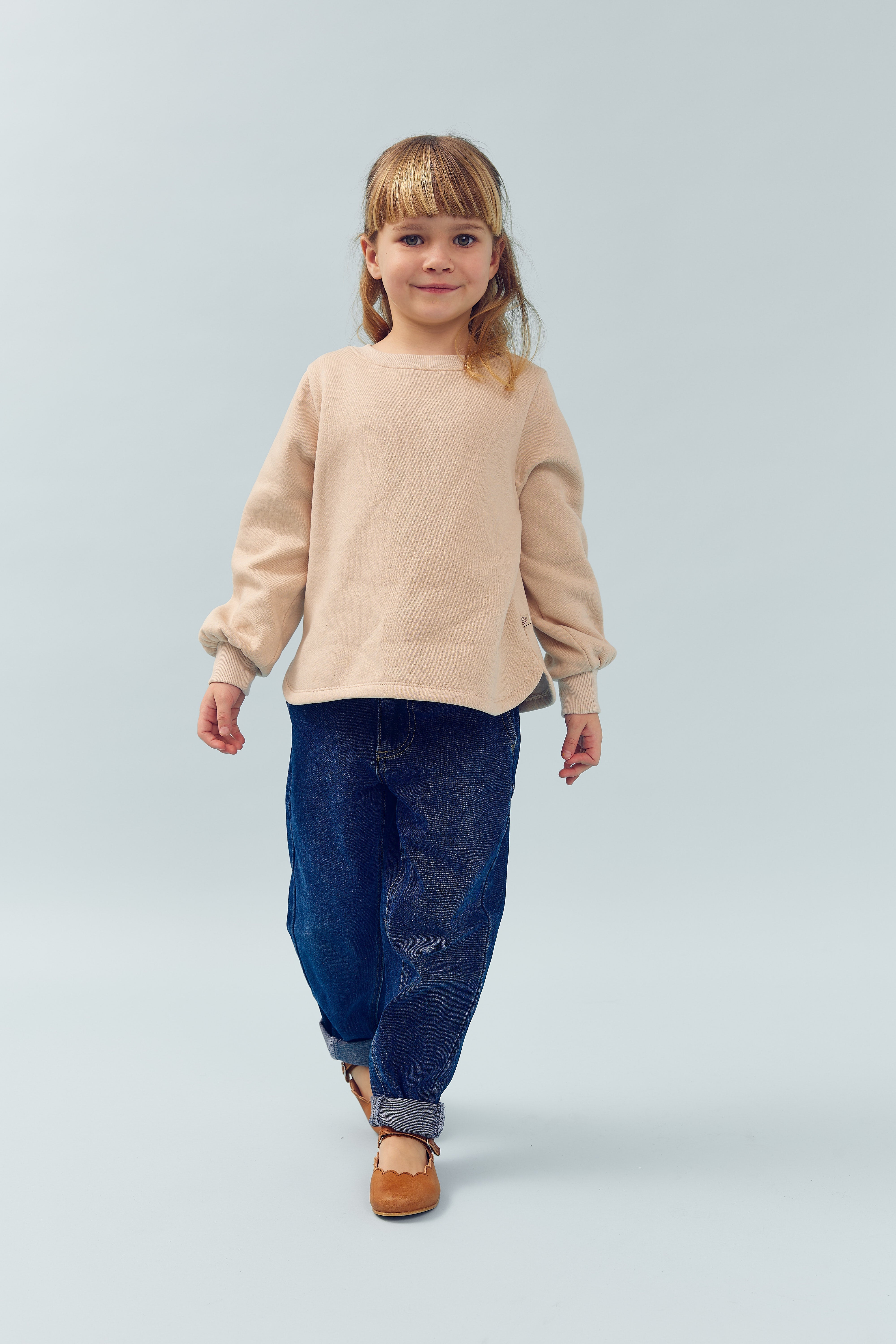 Et barn iført beige trøje og blå jeans går fremad. Baggrunden er enkel og lys grå, hvilket giver et rent og minimalistisk udtryk.