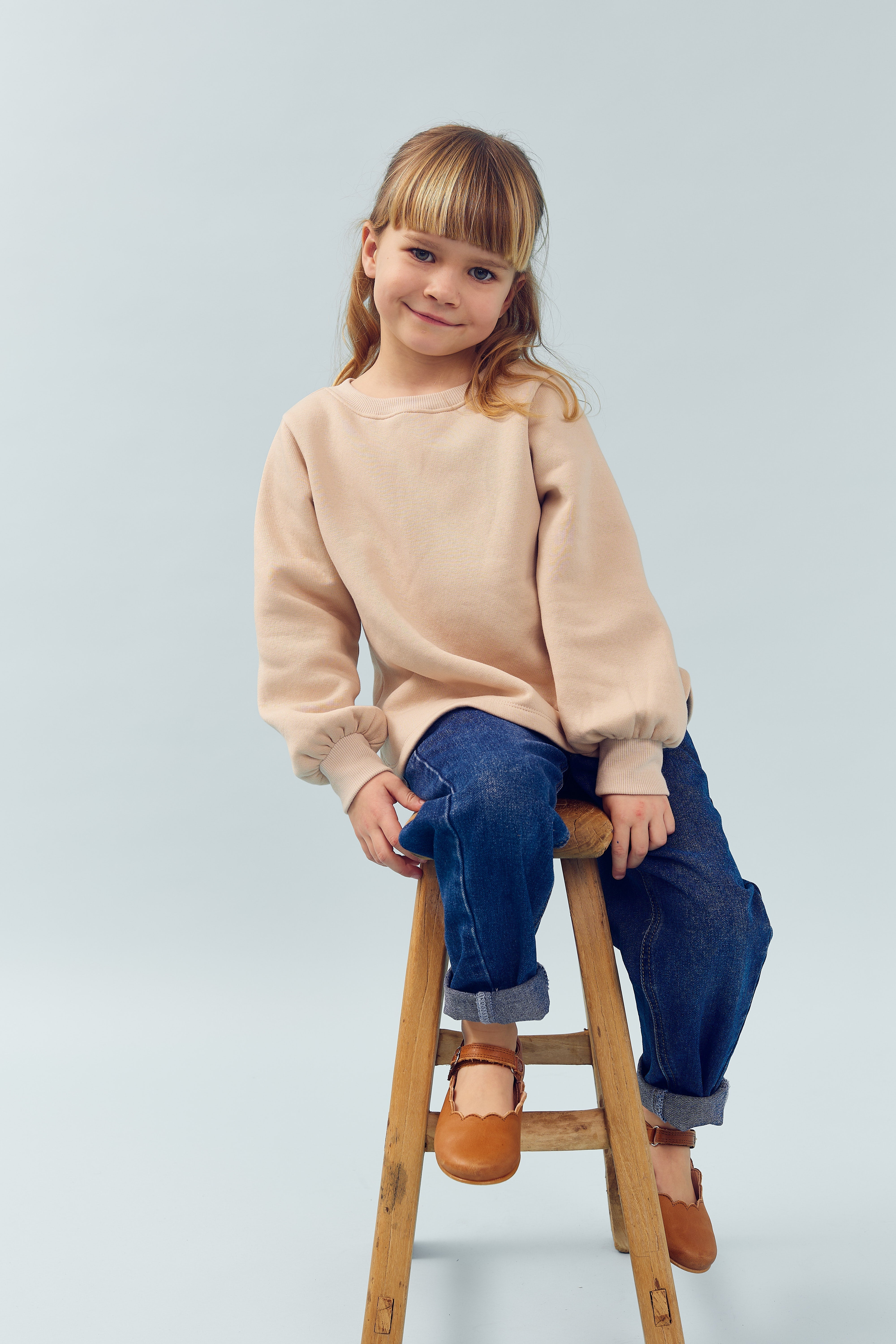 Et barn sidder afslappet på en træstol iført en beige trøje og blå jeans. Barnet smiler blidt i et neutralt, lysblåt studieomgivelse.