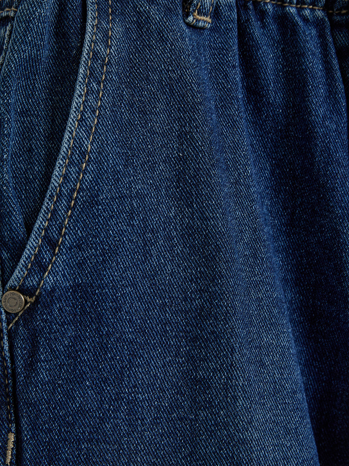 Denimstof vises tæt på, afslører en mørkeblå farve og tydelige syninger. Stoffet har en robust tekstur, typisk for jeans, med en lille del af en metalknap synlig i hjørnet.