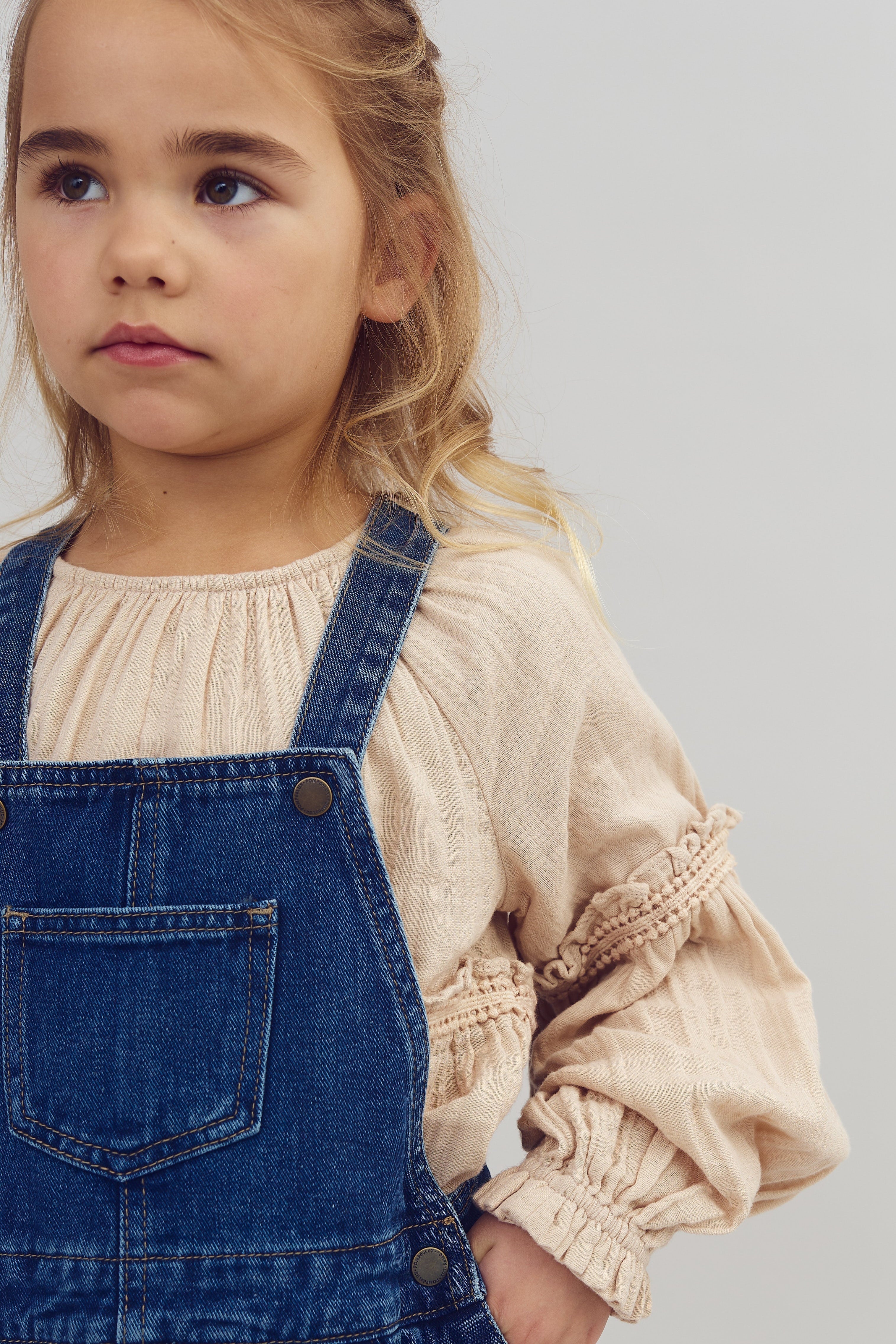 Et barn kigger til siden, iført en beige bluse og blå denim overall, står imod en lys baggrund.