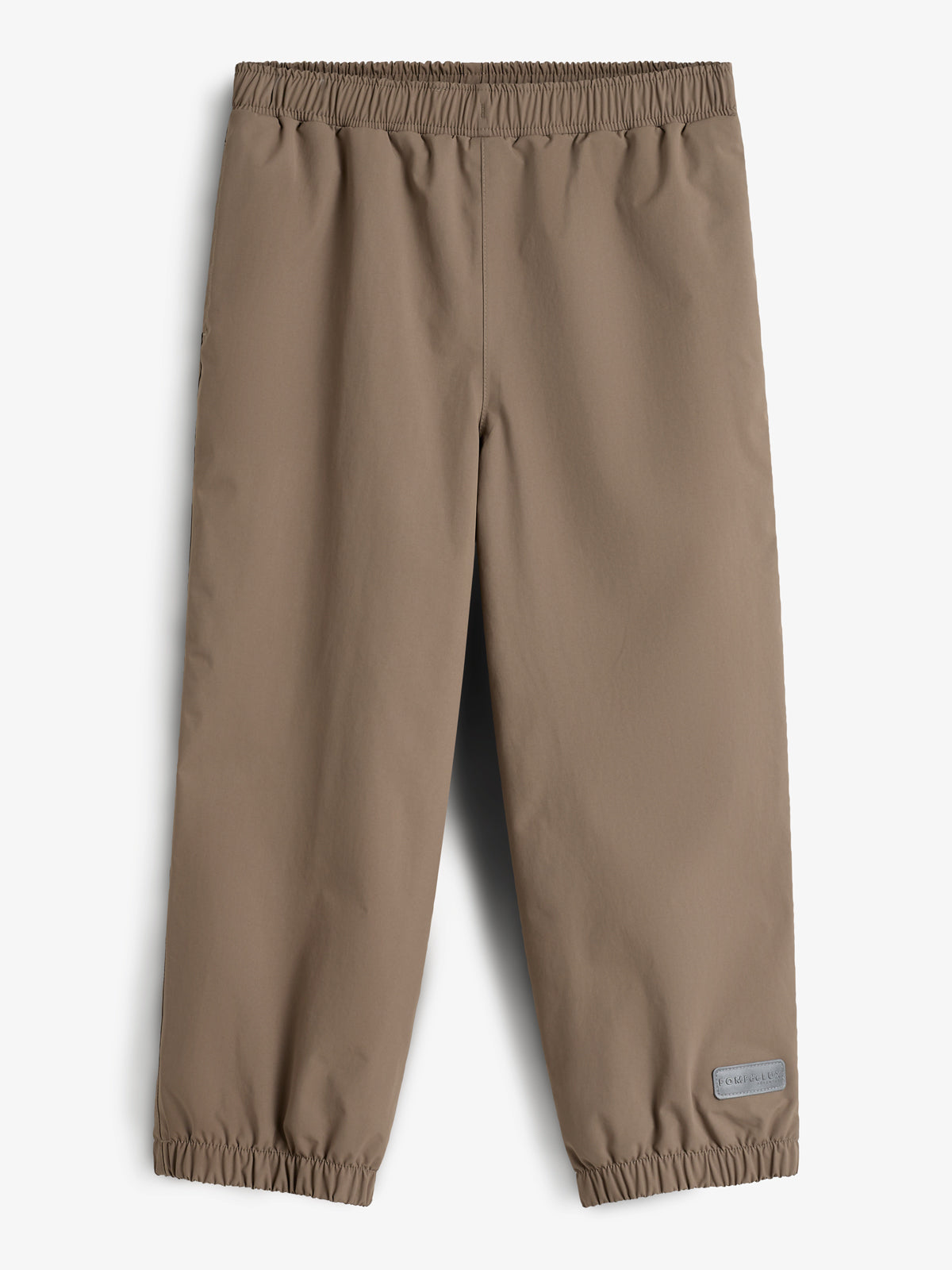 Bukser med elastisk talje og ankler, udført i en beige farve. Et lille rektangulært mærke nederst på det ene ben. Baggrunden er neutral hvid.