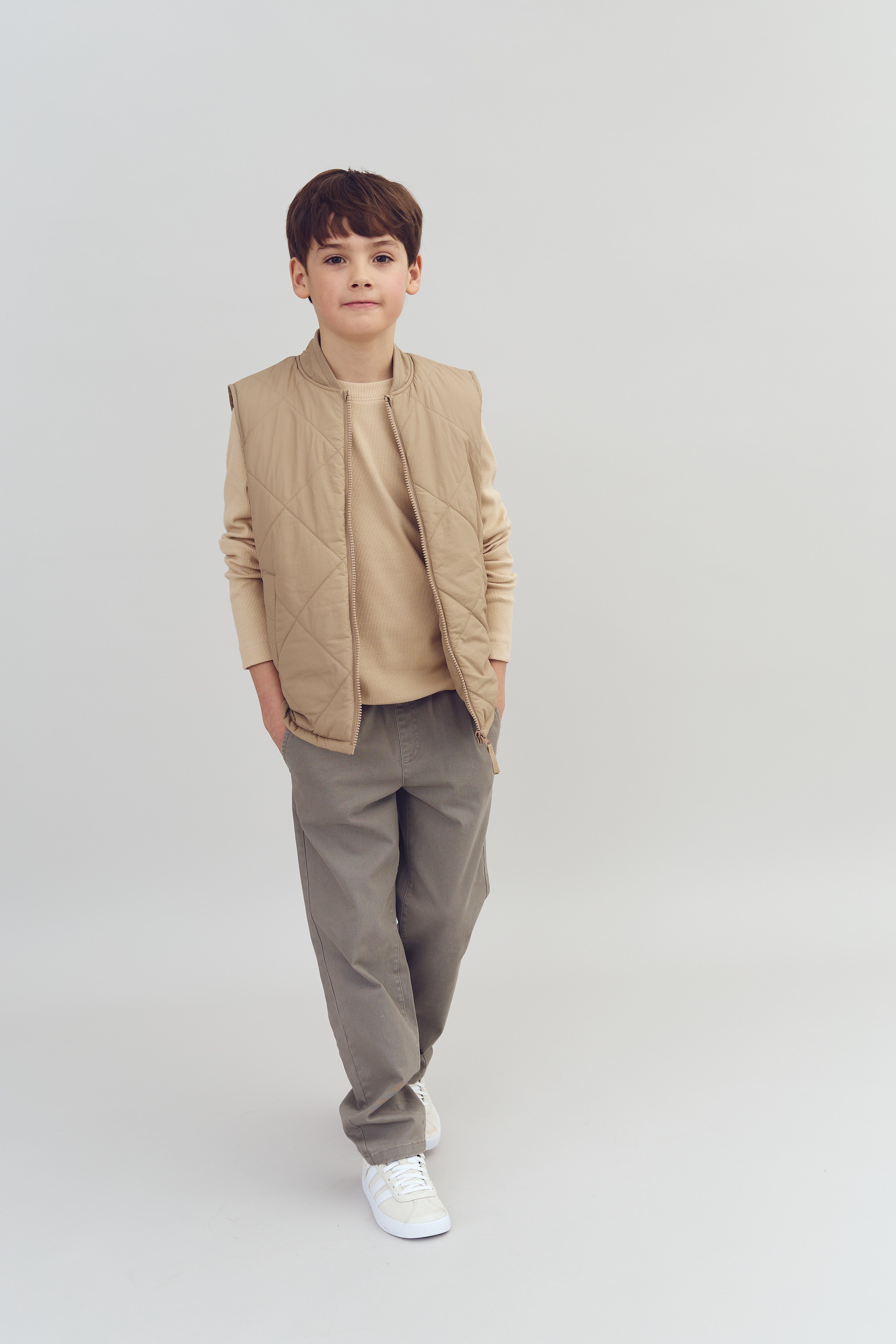 Dreng i beige vest står med hænderne i lommerne i lyse bukser og sneakers. Baggrunden er neutral og lys, hvilket fremhæver hans afslappede stil.