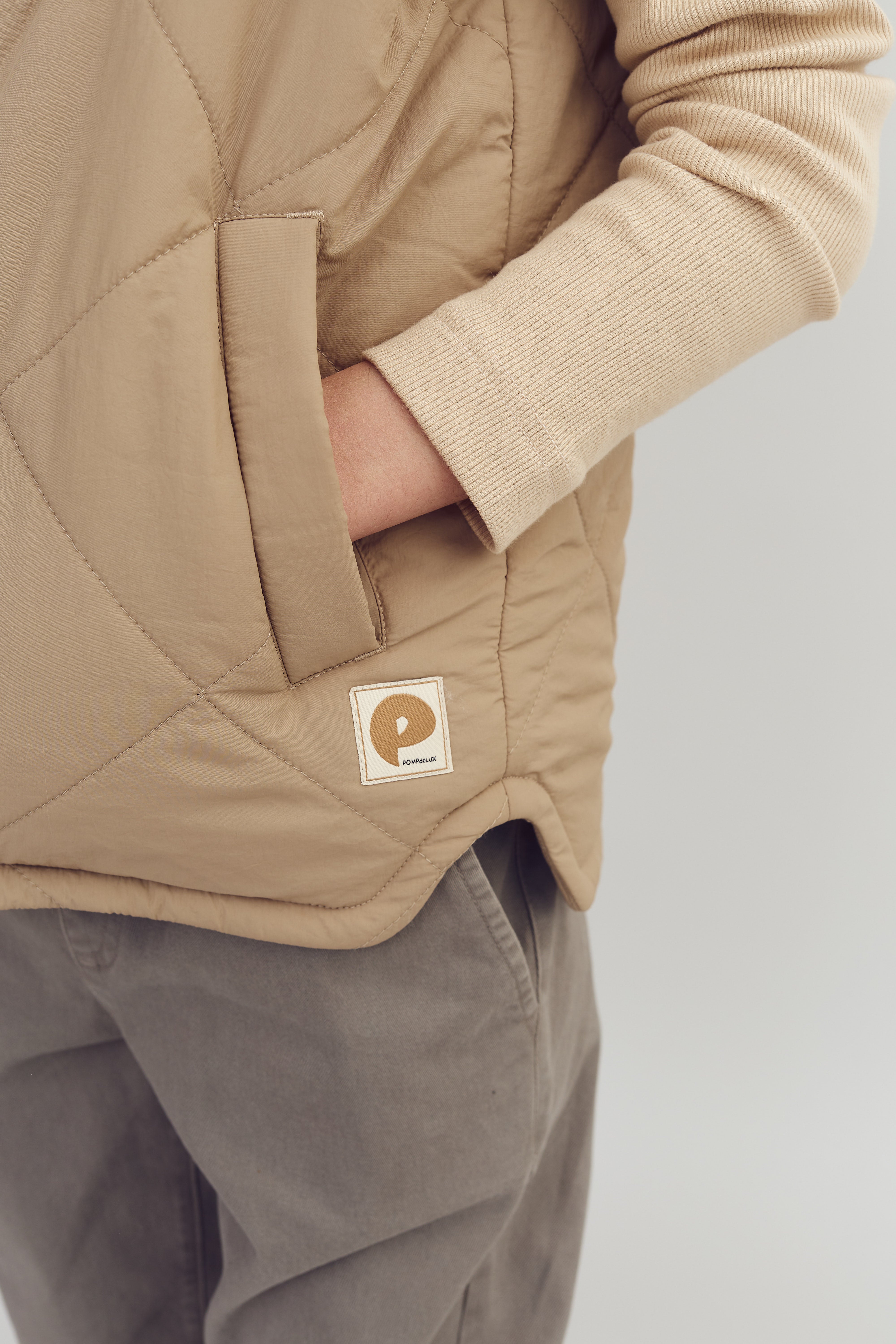 En person iført en beige jakke med hånden i lommen. Jakken har en mærkat med teksten "POPMAKINS." Personen bærer også grå bukser, set mod en lys baggrund.