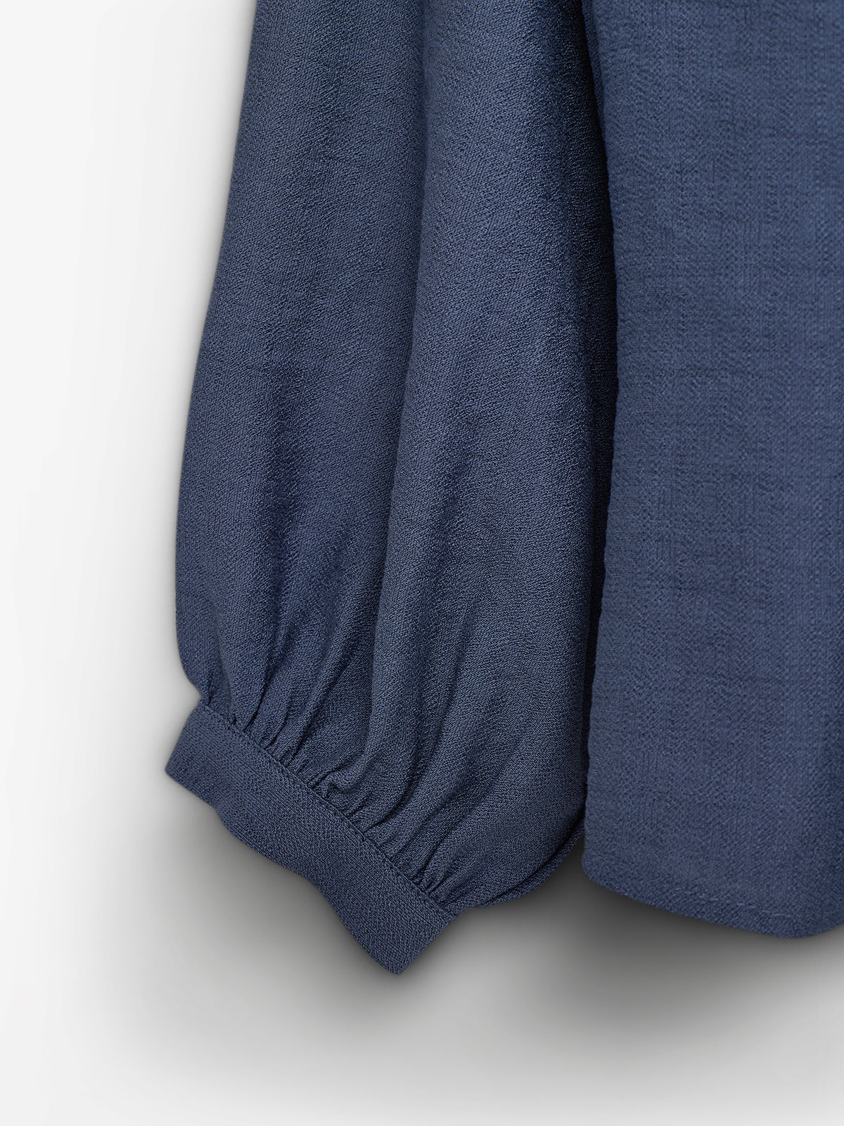 Blå bluseærme, der hænger løst ned. Ærmet har en elastisk kant, der samler stoffet ved håndleddet. Baggrunden er enkel og lys.
